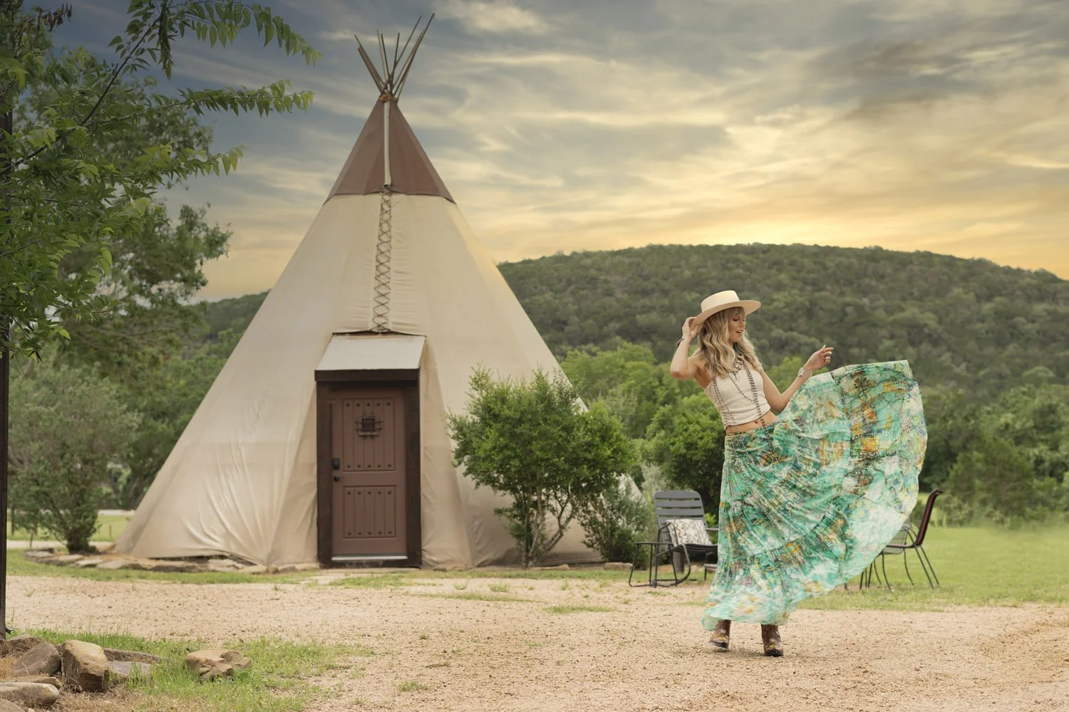 Woman wearing long flowy skirt and cowboy hat, posing in front of a teepee