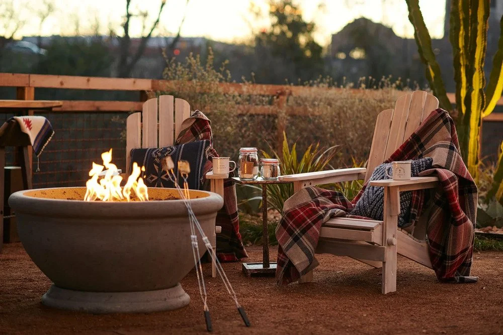  Outdoor fire pit surrounded by wooden Adirondack chairs, plaid blankets, and mugs at sunset 