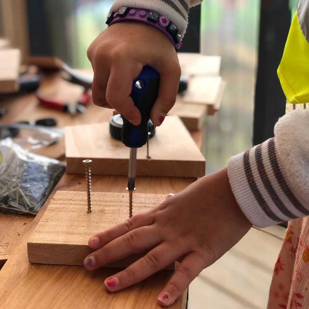 Child using a stubby screwdriver to fasten a screw into wood in a preschool woodworking session