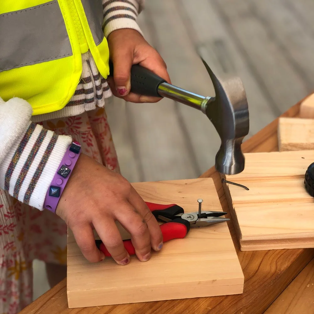 Child holding a nail with pliers while hammering wood in a supervised classroom carpentry session