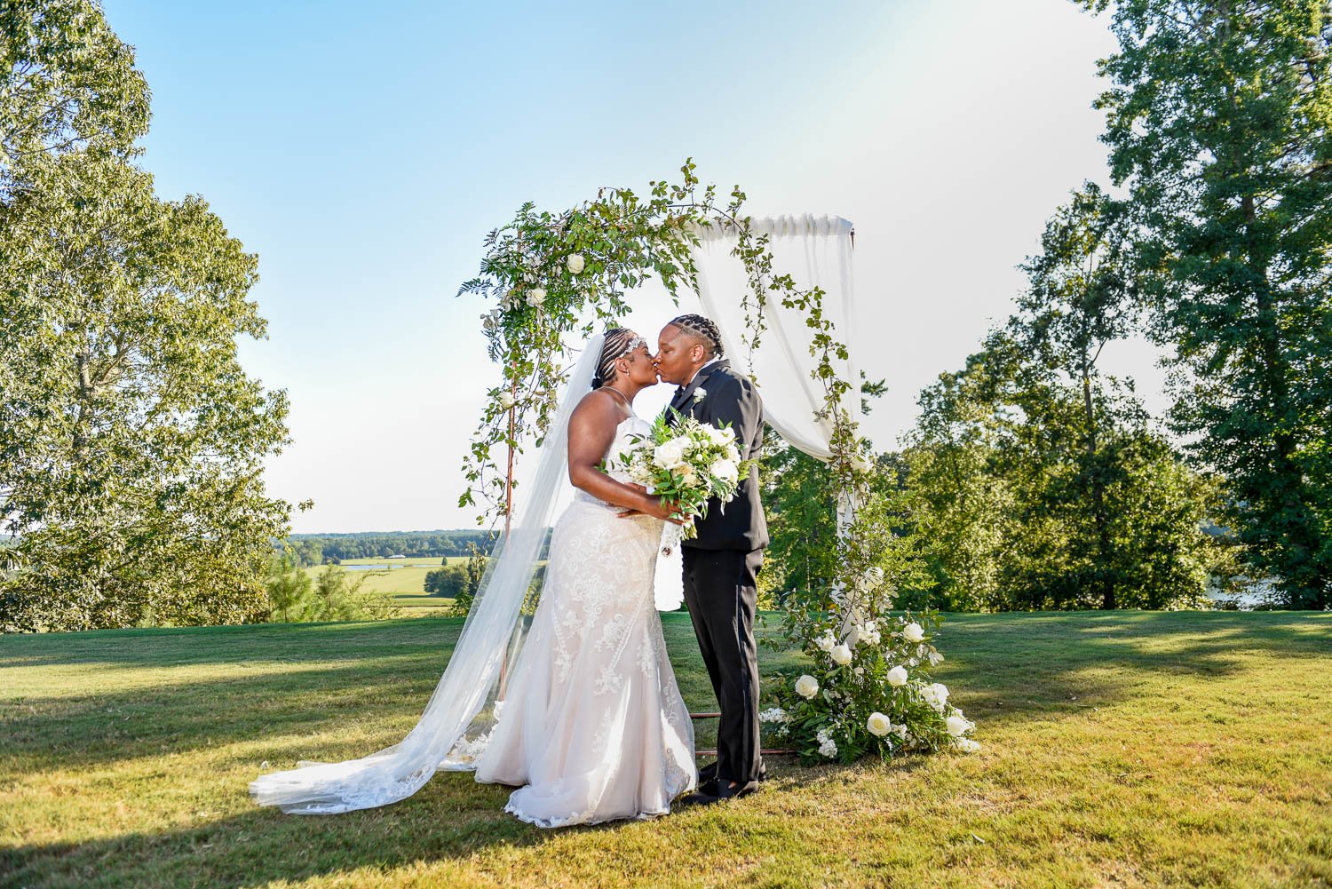 Brides Candace and Mecha Pose in front of their floral arch atop Foxhall Resorts highest point for wedding day portraits and seal it with a kiss
