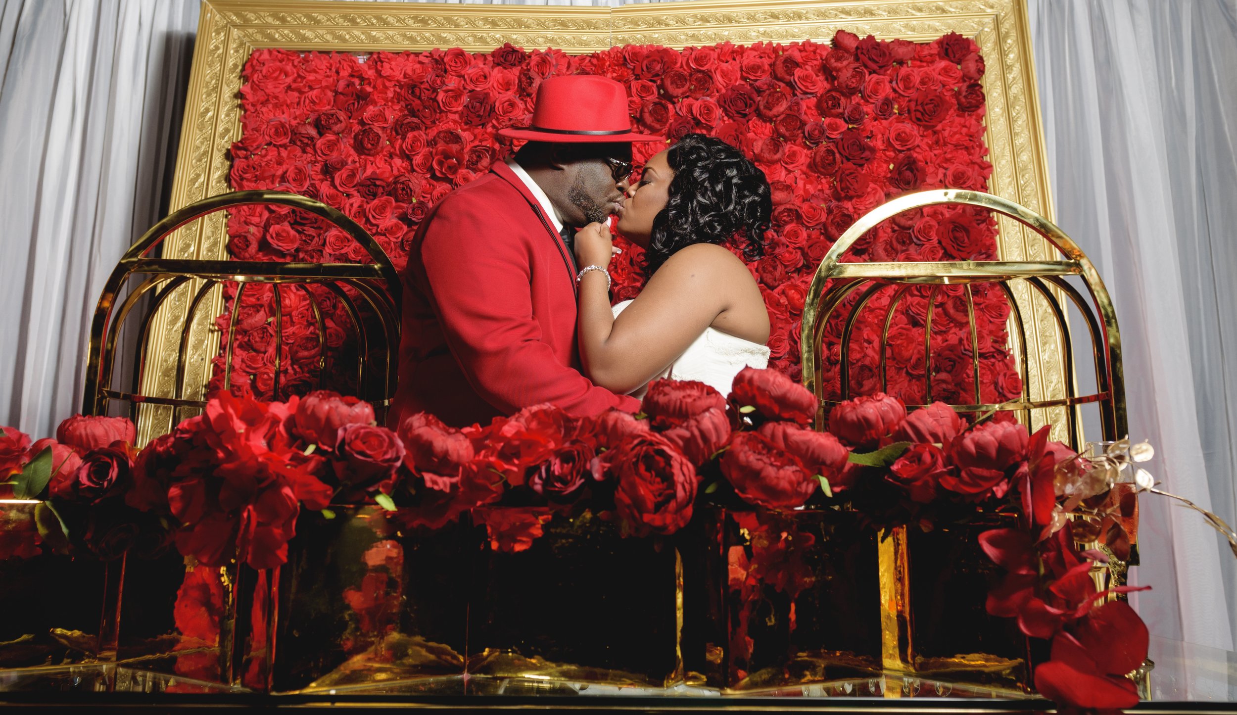 Bride and groom stand in front of their Red rose filled flower wall to capture a moment in front of their gold throne chairs during their wedding reception