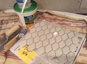 Supplies for installing the tile surrounding the fireplace insert; hexagonal mosaic tile, a small tub of premium mortar, and trowels in various sizes.