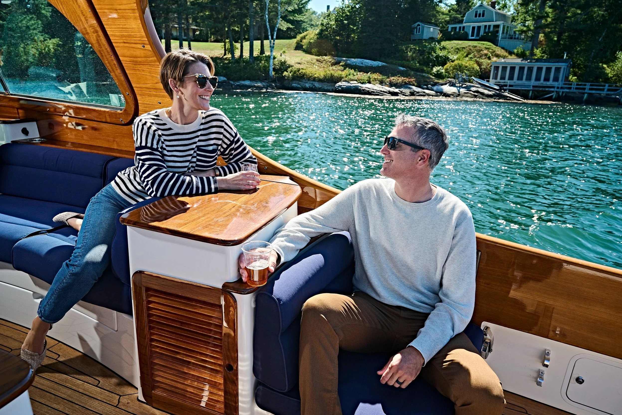 Two people relaxing and smiling on a boat with drinks, near a scenic shoreline.