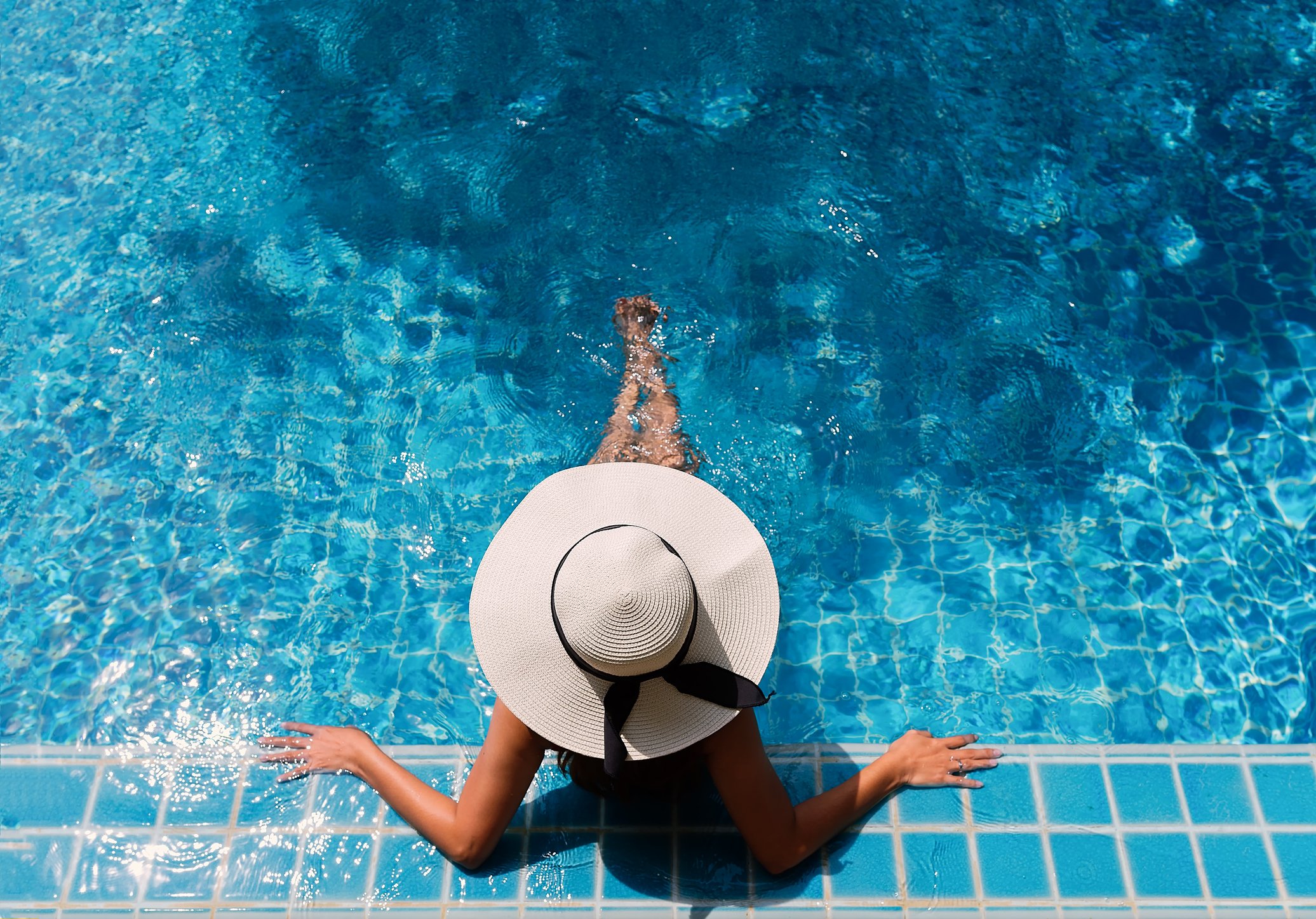 A woman in a black swimsuit and a wide-brimmed straw hat with a black ribbon is sitting on the edge of a swimming pool, with her feet in the water and her arms resting on the pool edge. The pool has blue tiles and the water is clear and blue.