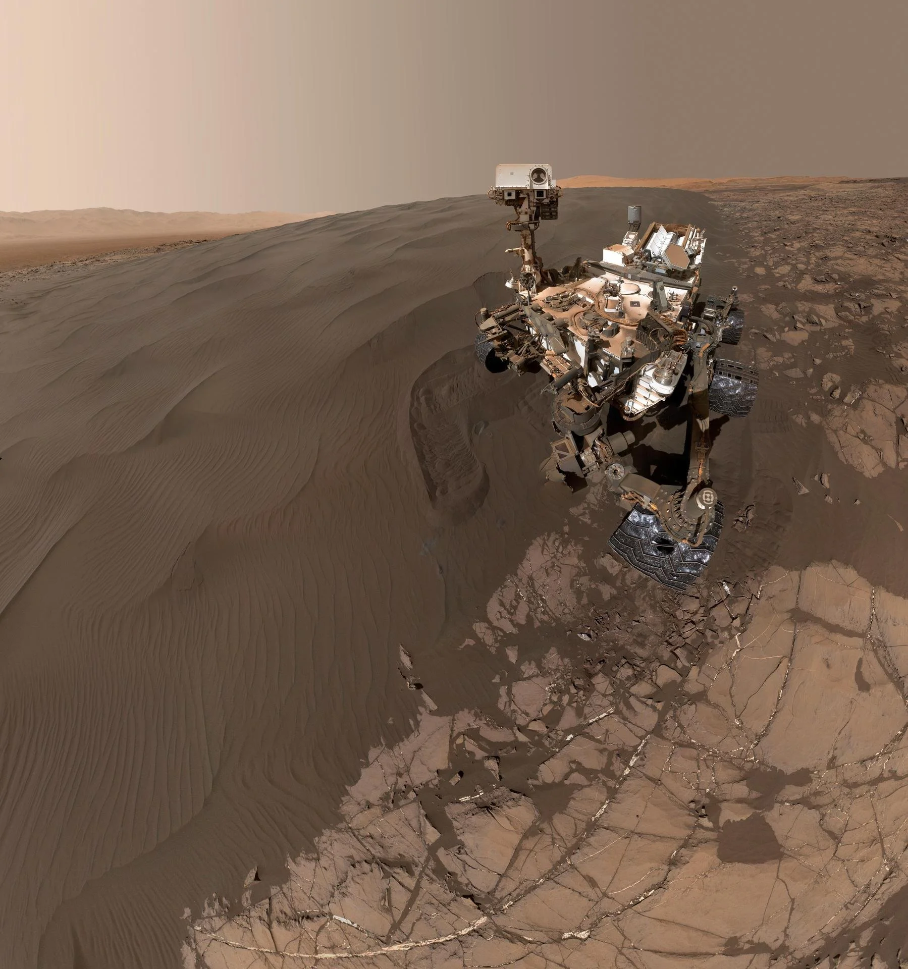 Curiosity rover self-portrait beside a dark Martian sand dune, showing its robotic arm, wheels, and instruments against rippled dunes and cracked bedrock under a hazy Mars sky.