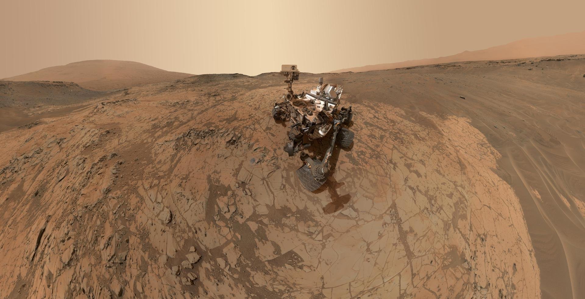 Self-portrait of NASA’s Curiosity rover on Mount Sharp, showing the rover on fractured bedrock with sandy slopes and distant hills under a dusty Martian sky.