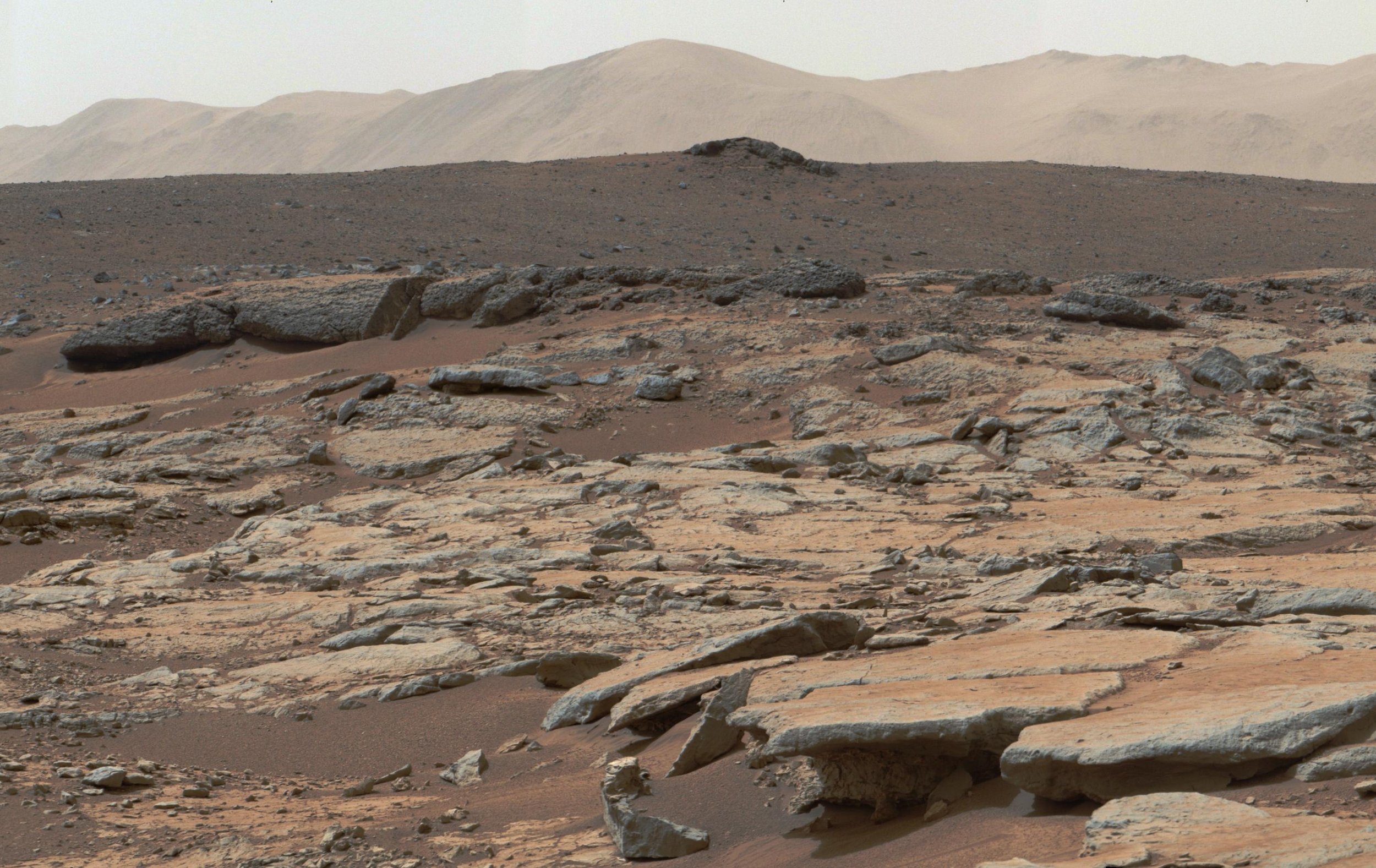 Mastcam mosaic from NASA’s Curiosity rover showing layered sedimentary rocks in Yellowknife Bay, Gale Crater, with flat slabs, scattered stones, and distant low hills under a hazy sky.