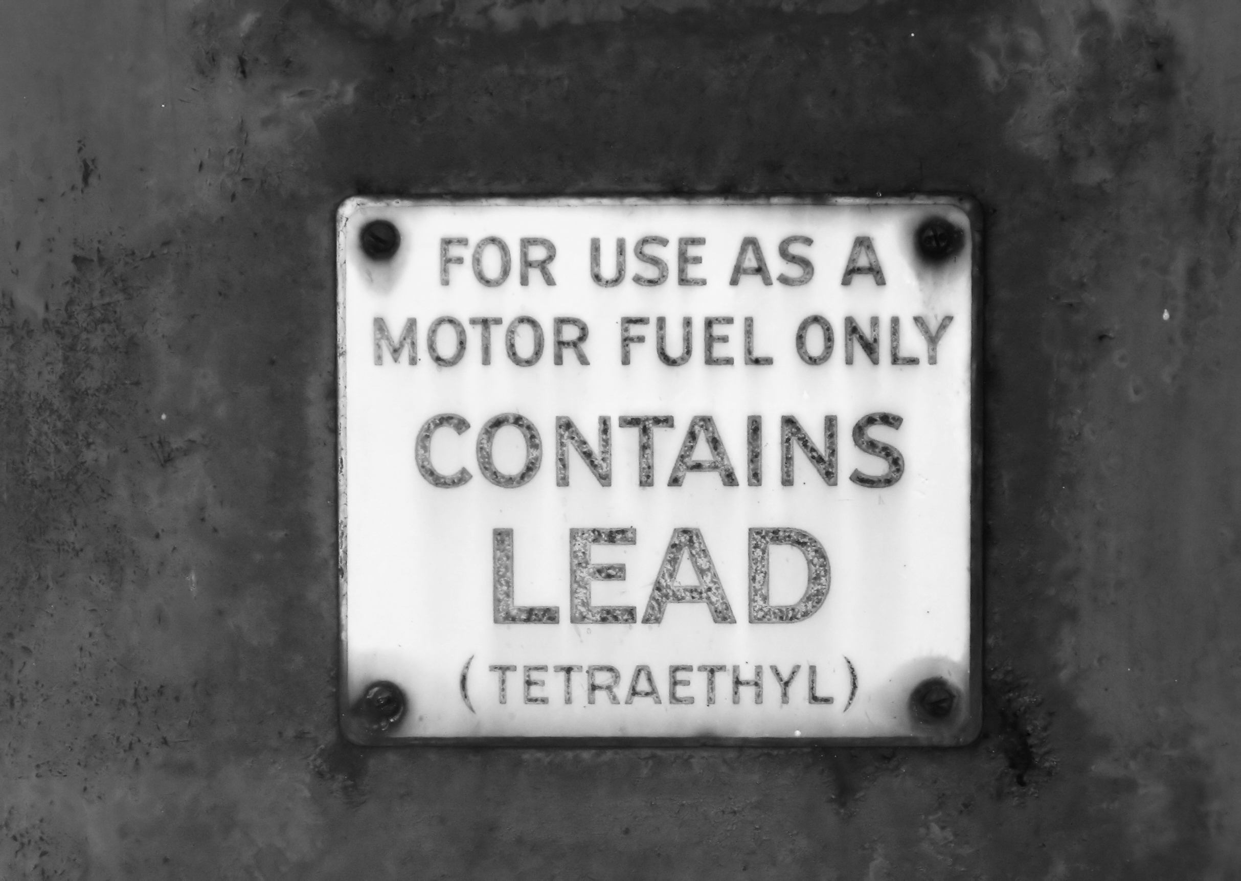 Black-and-white photograph of a metal sign reading "For Use as a Motor Fuel Only. Contains Lead (Tetraethyl)," mounted on a fuel tank or pump.