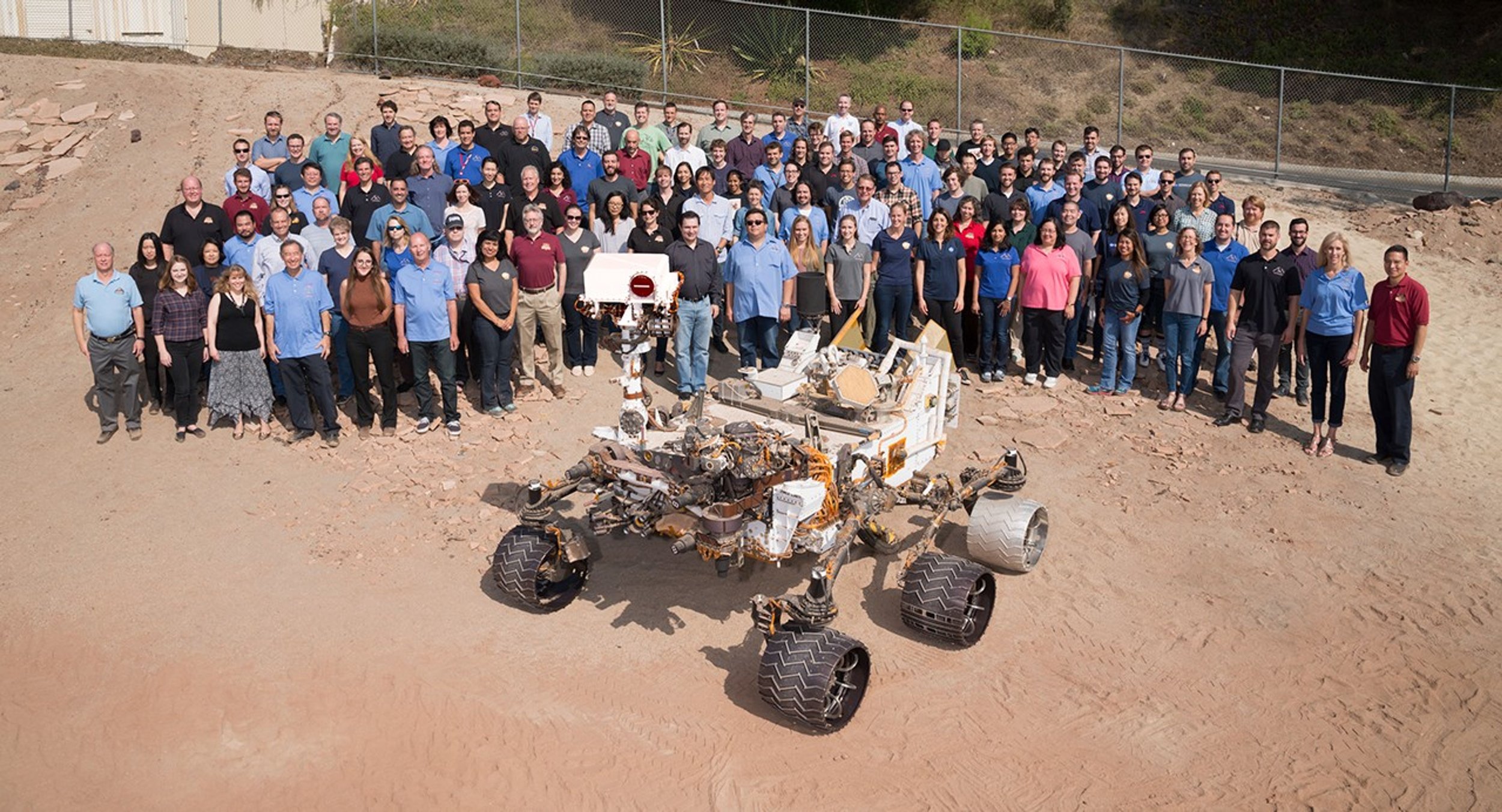 Group photo of the Mars Science Laboratory team standing behind a Curiosity test rover in JPL’s Mars Yard, a sandy outdoor test area.