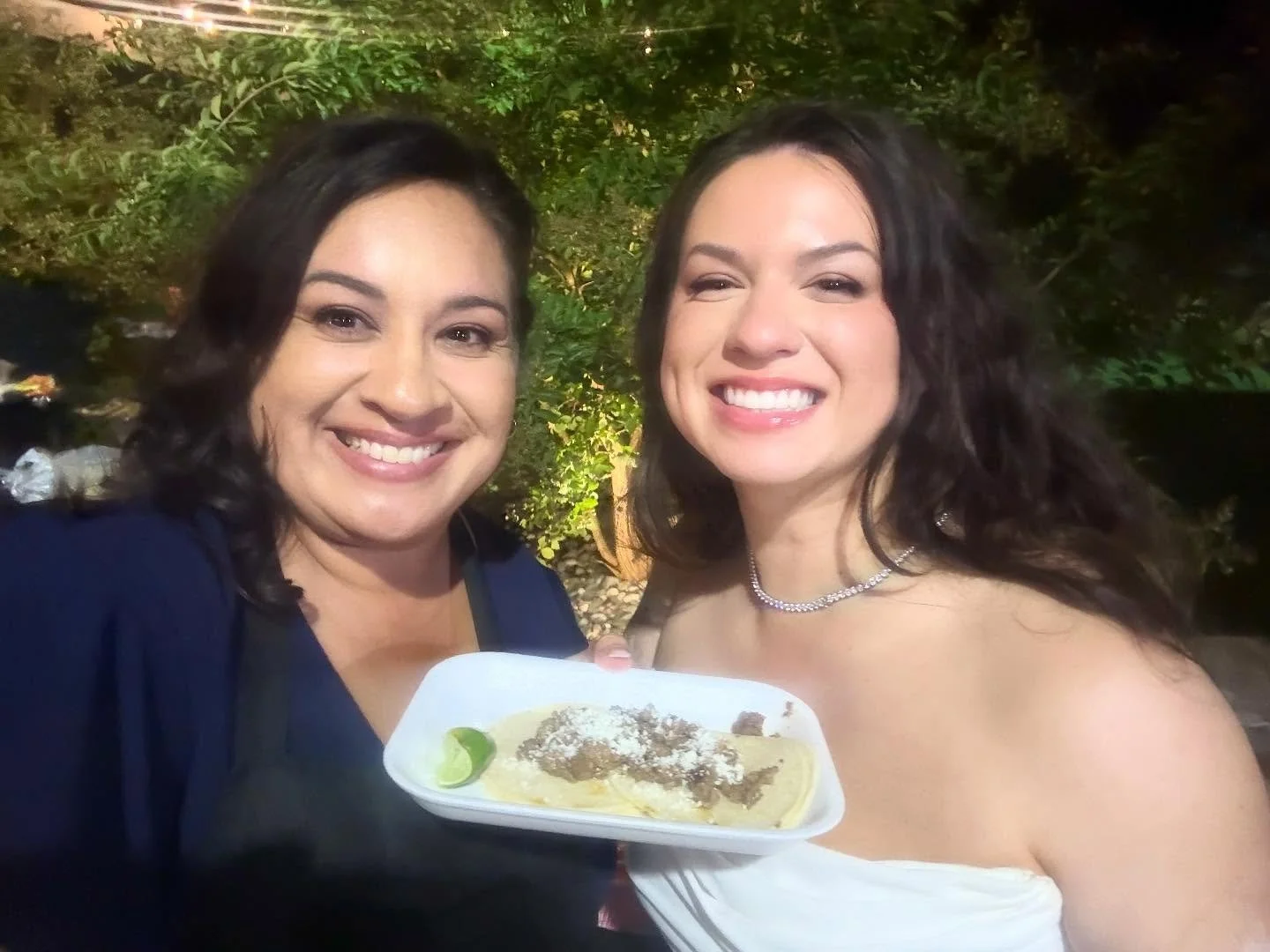 Two women smiling in an outdoor setting, one holding a plate of food that appears to be a taco or similar dish with lime, cheese, and meat, at night with greenery in the background.