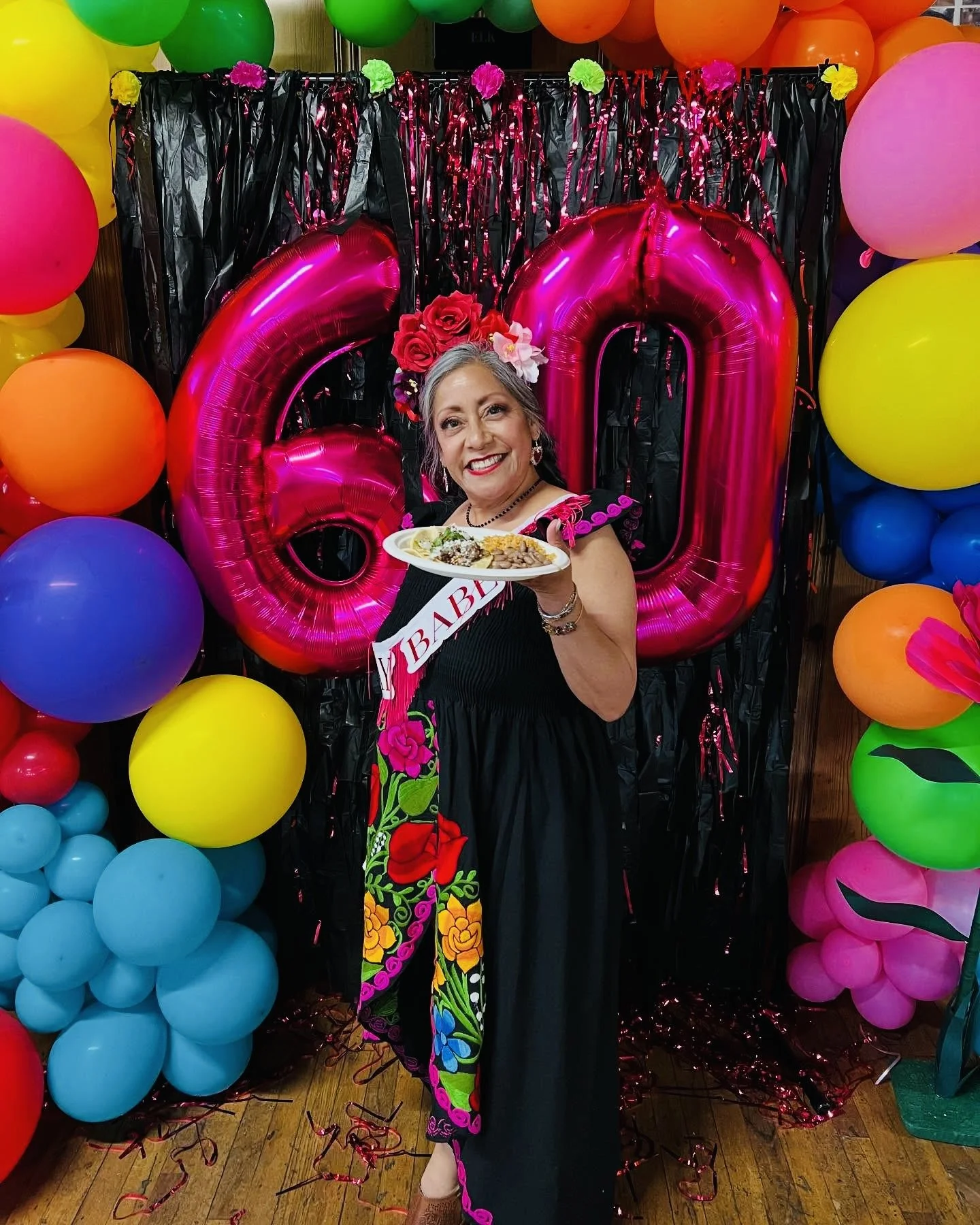 A woman at a celebration holding a plate of food, standing in front of colorful balloons, with a large pink balloon-digital '60' behind her, wearing a floral sash and crown, smiling at the camera.