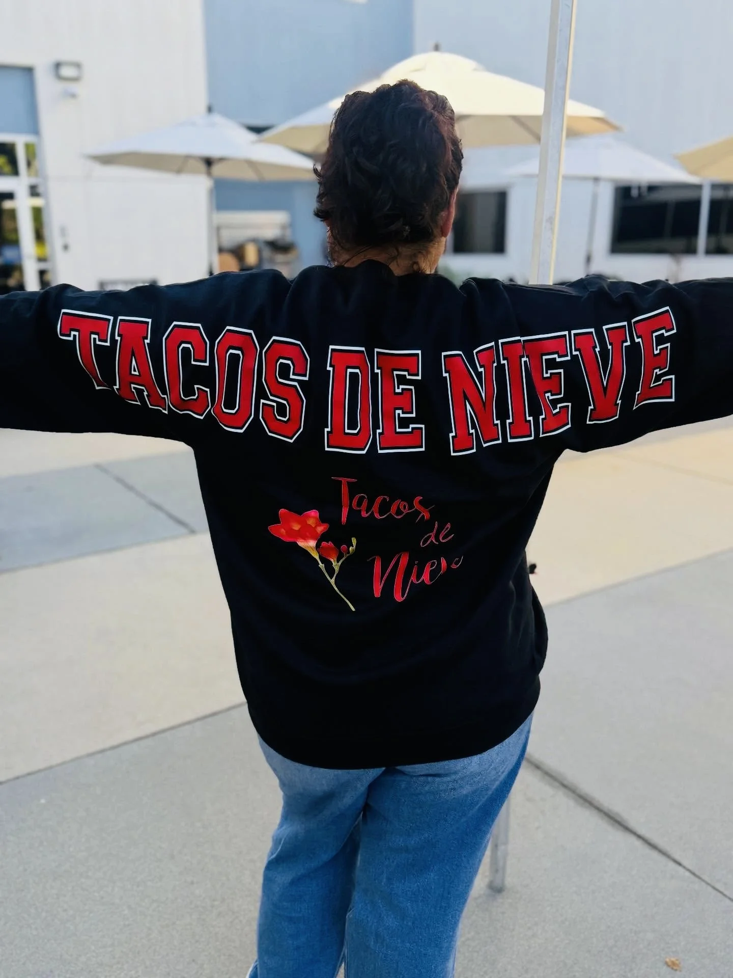 Person standing outside with arms outstretched, wearing a black jacket with red and white text that reads "TACOS DE NIEVE" and a flower graphic below, with umbrellas and a building in the background.