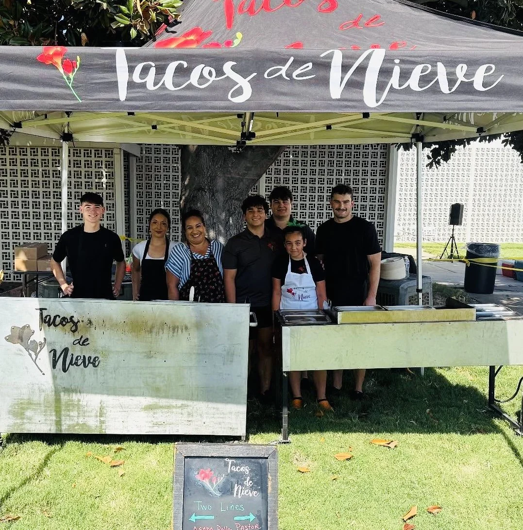 Group of seven people standing behind a food stand labeled "Tacos de Nieve," under a canopy with the same name. The stand is outdoors with a tree and a brick wall behind them, and a chalkboard sign in front with the menu and directions. The people ar
