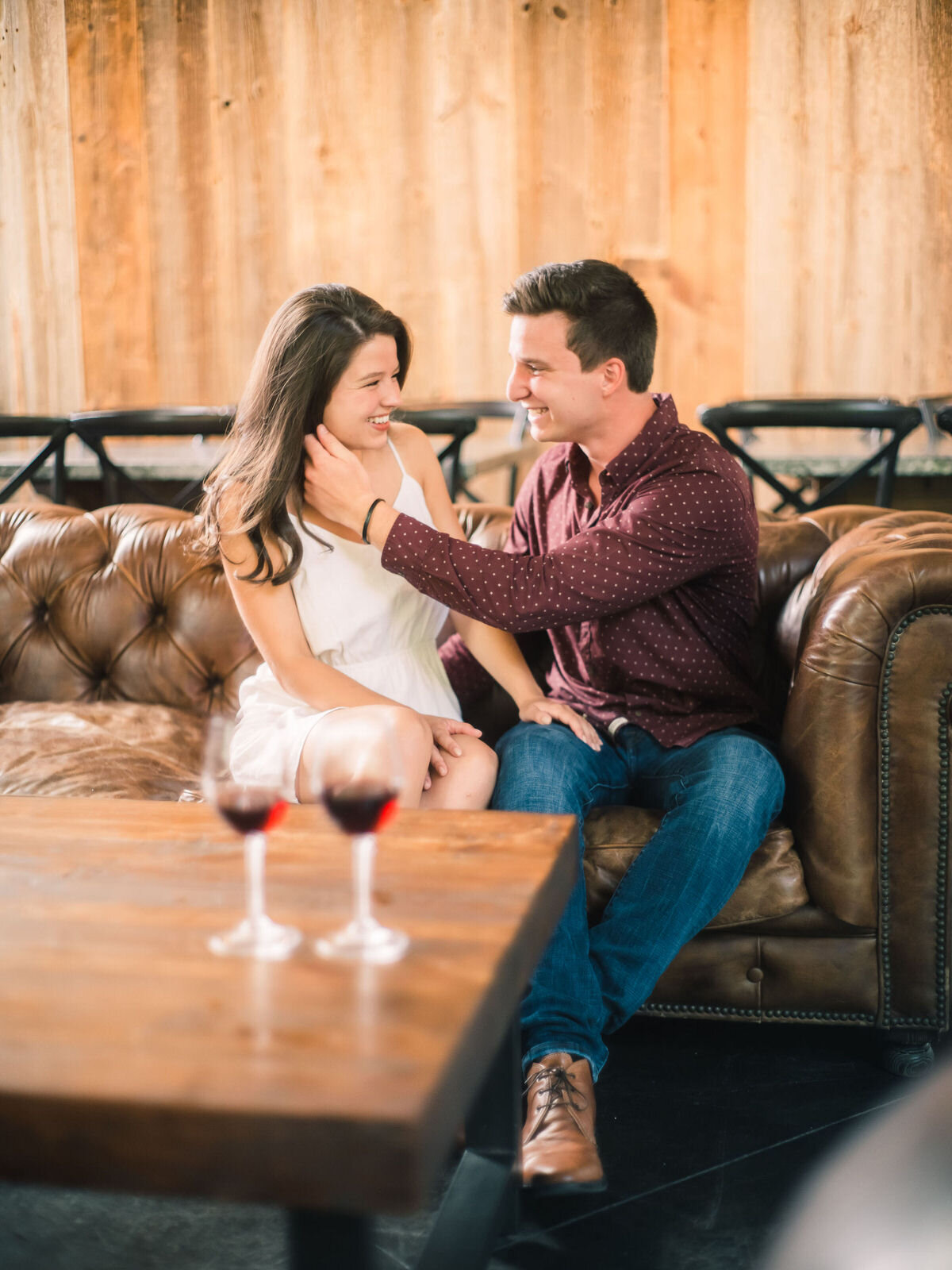 couple sitting on a couch with man reaching across to touch woman's face with wine in front of them smiling and laughing