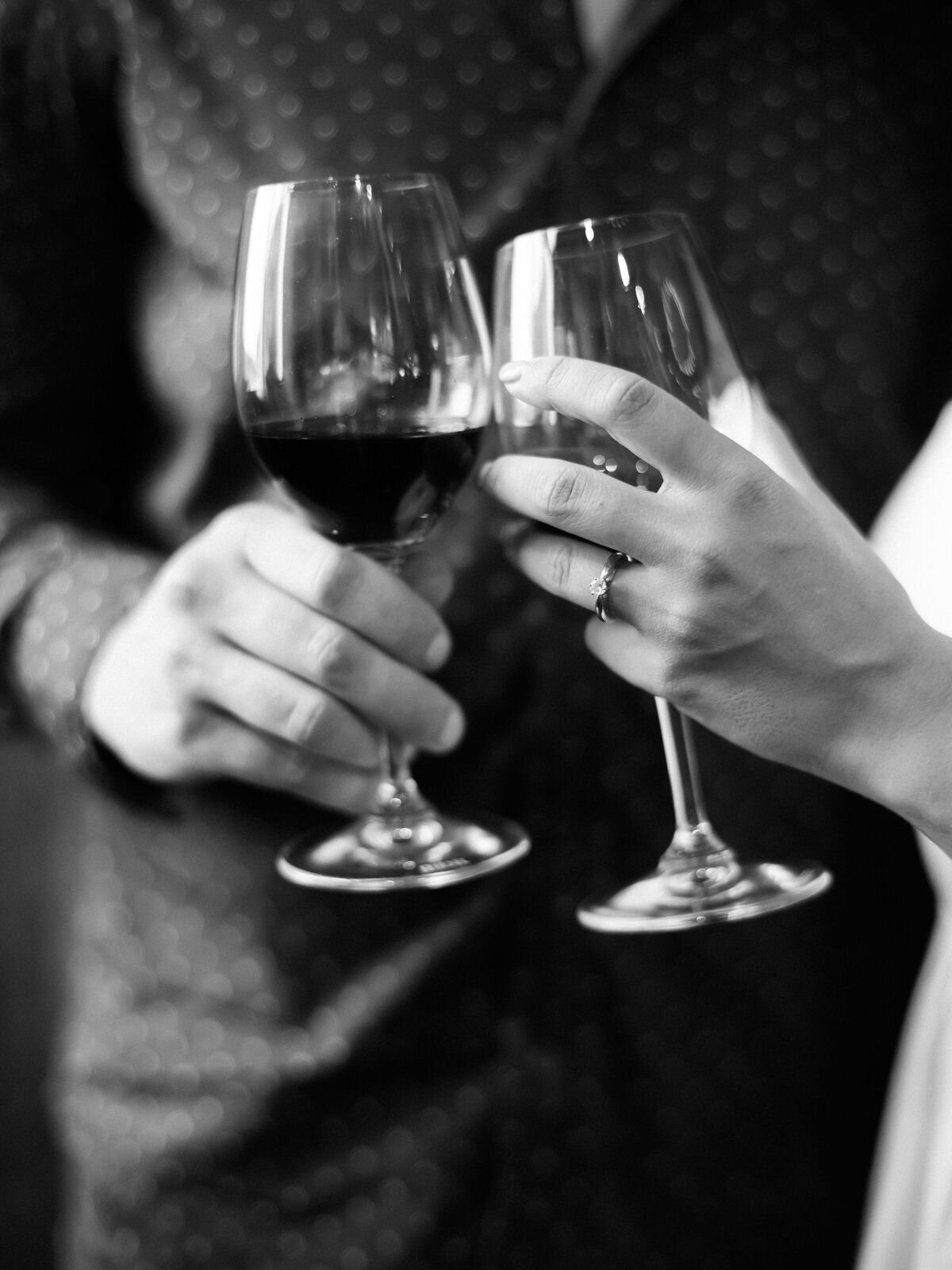 wine glasses in black and white with an engagement ring touching with a couple behind them toasting