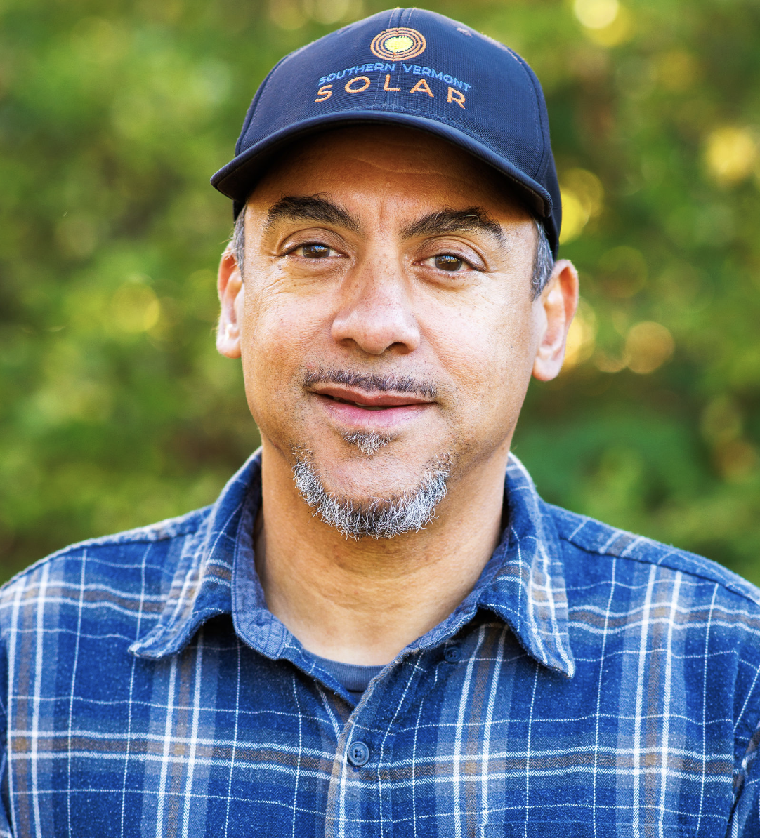 A middle-aged man wearing a blue baseball cap with a 'Southern Vermont Solar' logo and a blue plaid shirt, standing outdoors with green foliage in the background.