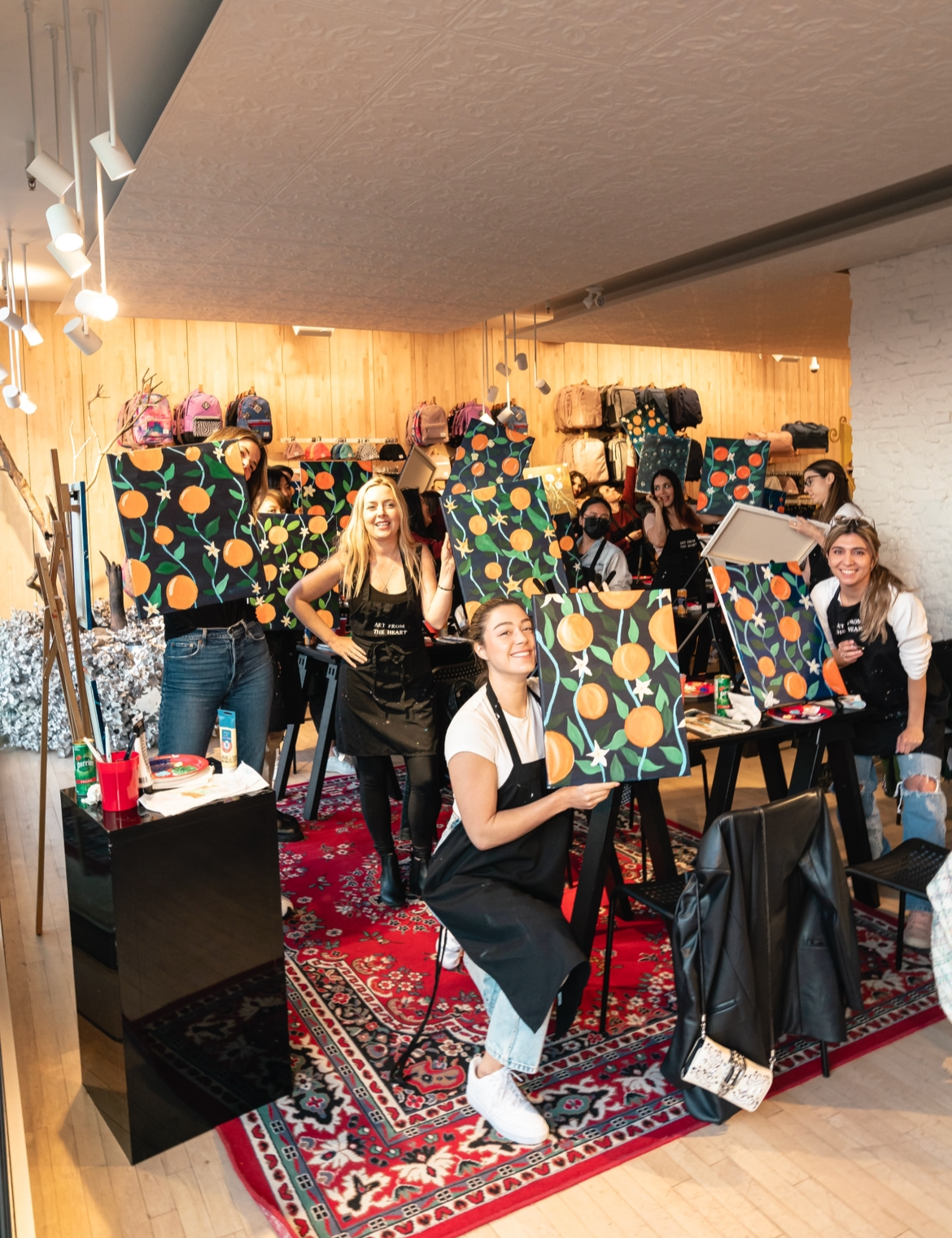 Group of people participating in a painting class, creating paintings of oranges and flowers on canvases. The room is decorated with a rug, wooden wall, and bags on shelves.
