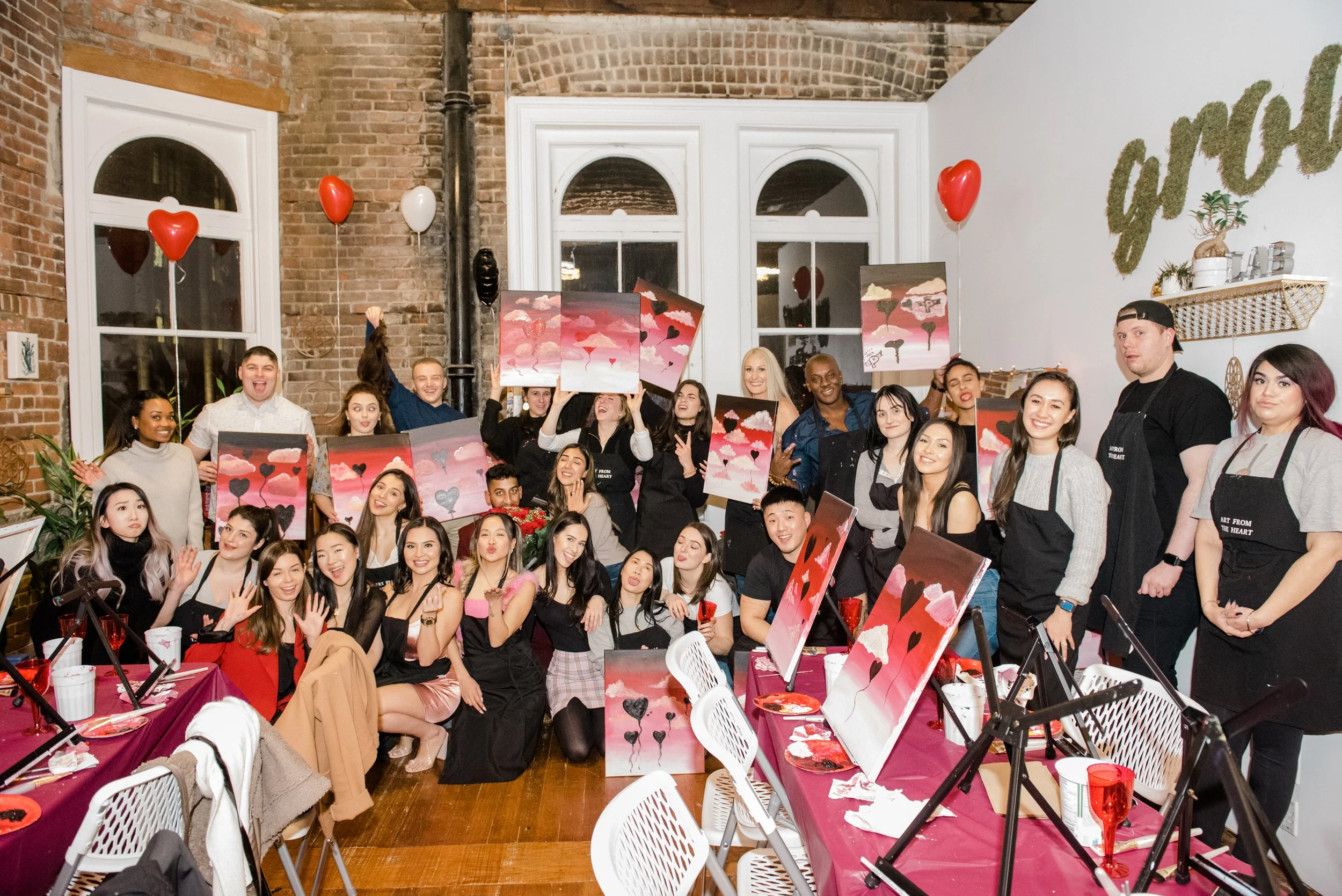 A group of people celebrating at an art party, displaying their paintings featuring hearts and pink clouds. The event has balloons, tables with art supplies and drinks, and a brick and white wall backdrop with the word 'love' on the wall.