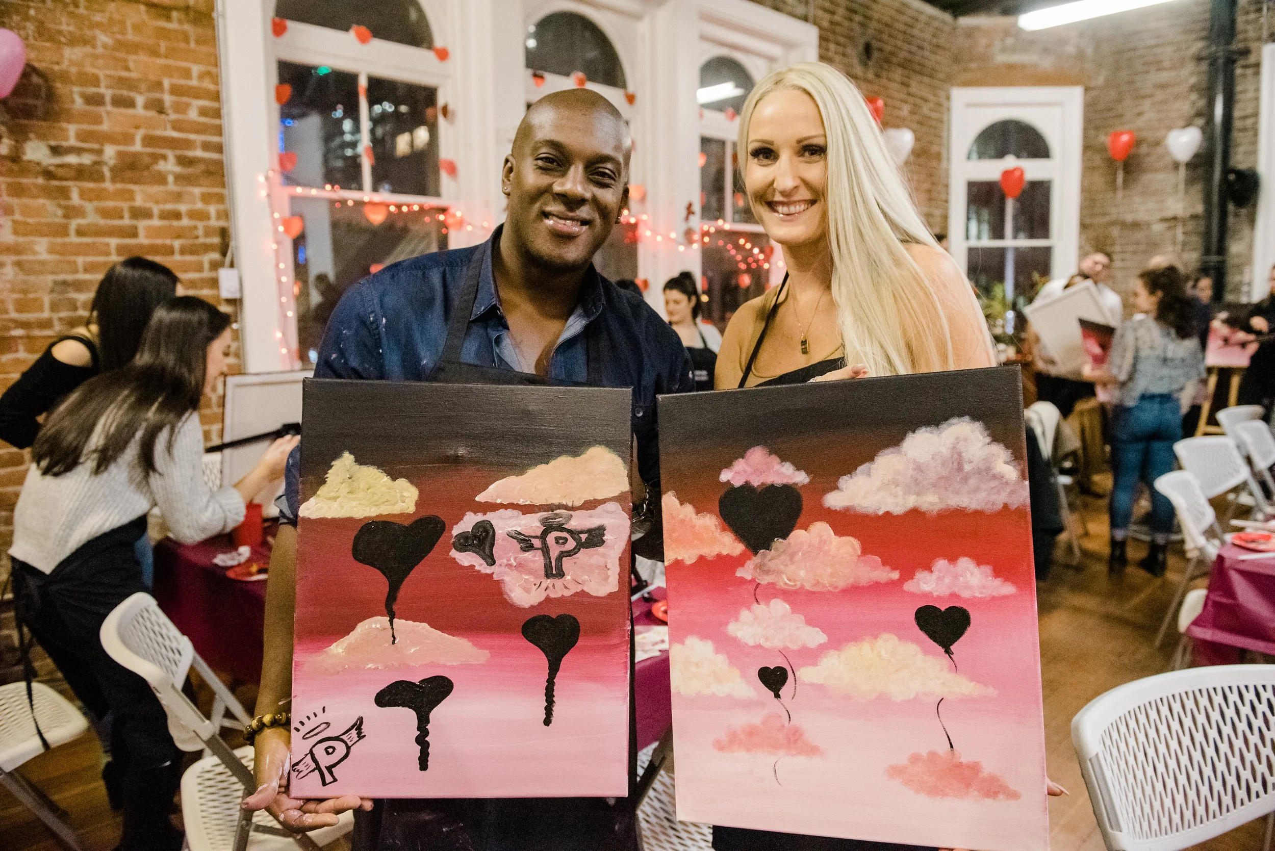 Two women holding paintings of clouds and hearts at a celebration event with Valentine's Day decor.