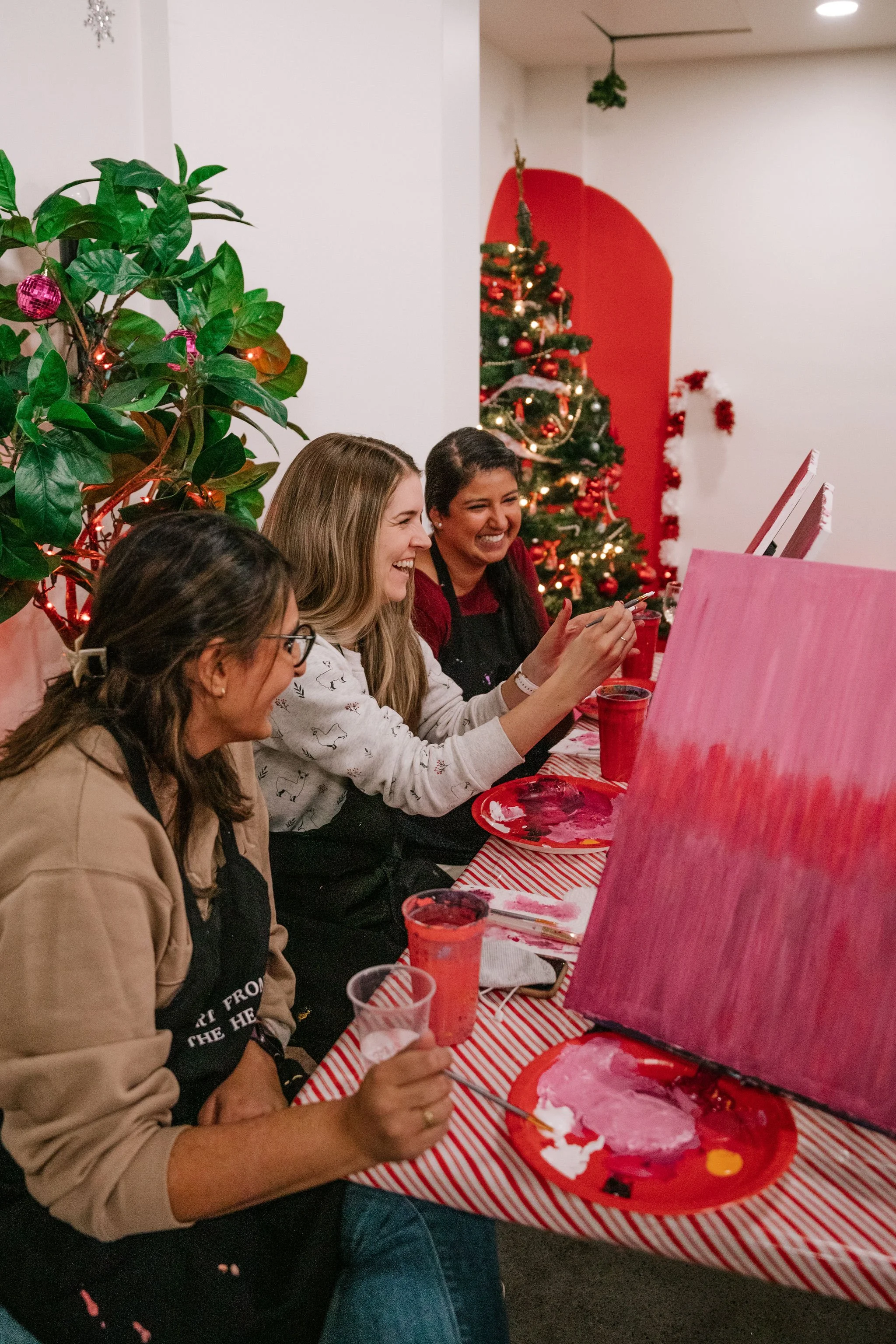 Several women sitting at a holiday-themed table, painting on canvases, with Christmas decorations including a Christmas tree and red ornaments in the background.