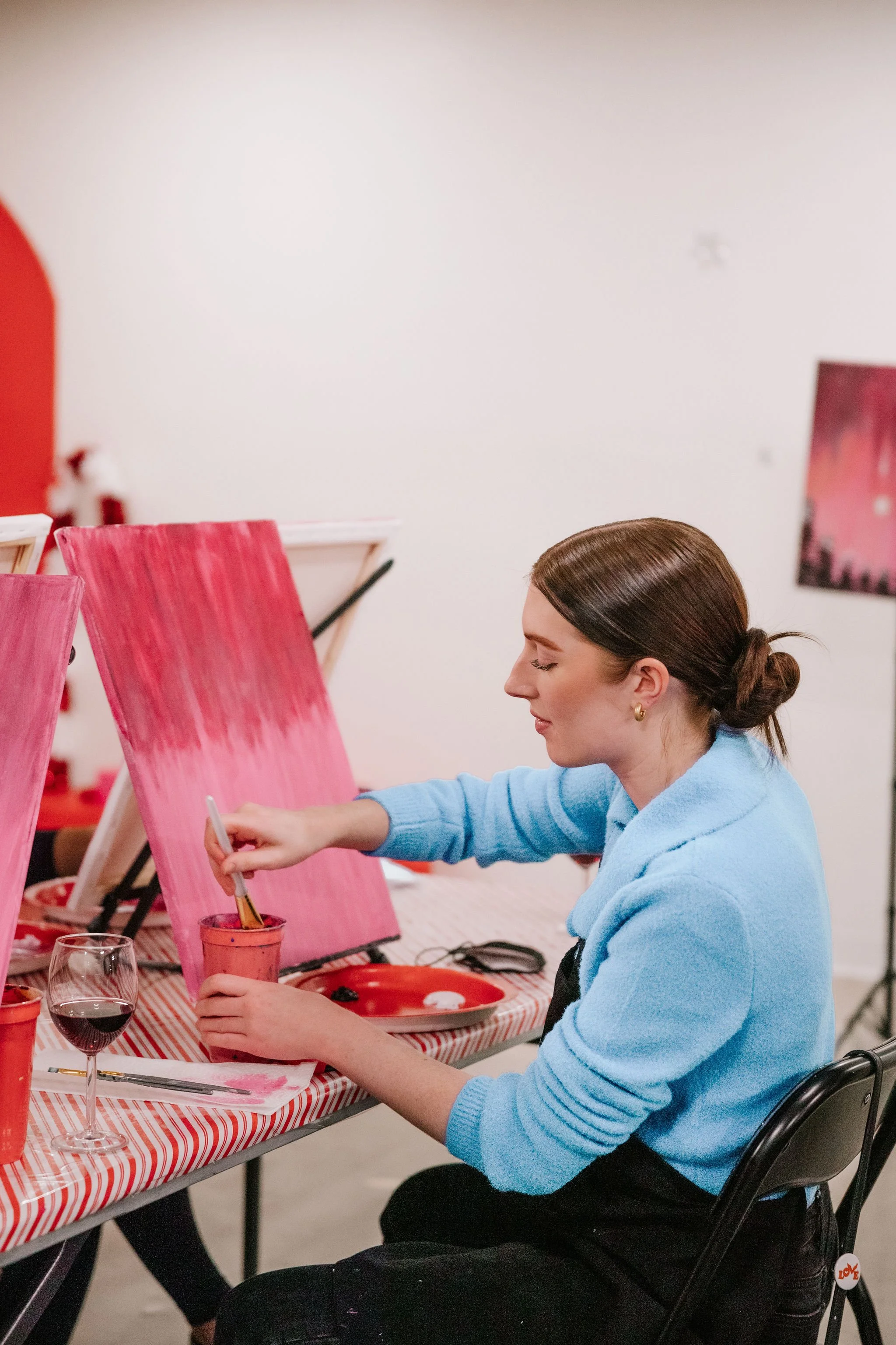 A woman with brown hair tied back in a bun, wearing gold hoop earrings and a light blue sweater, is sitting in a wheelchair at a table painting on a pink canvas. There are painting supplies, a wine glass, and a striped tablecloth on the table.
