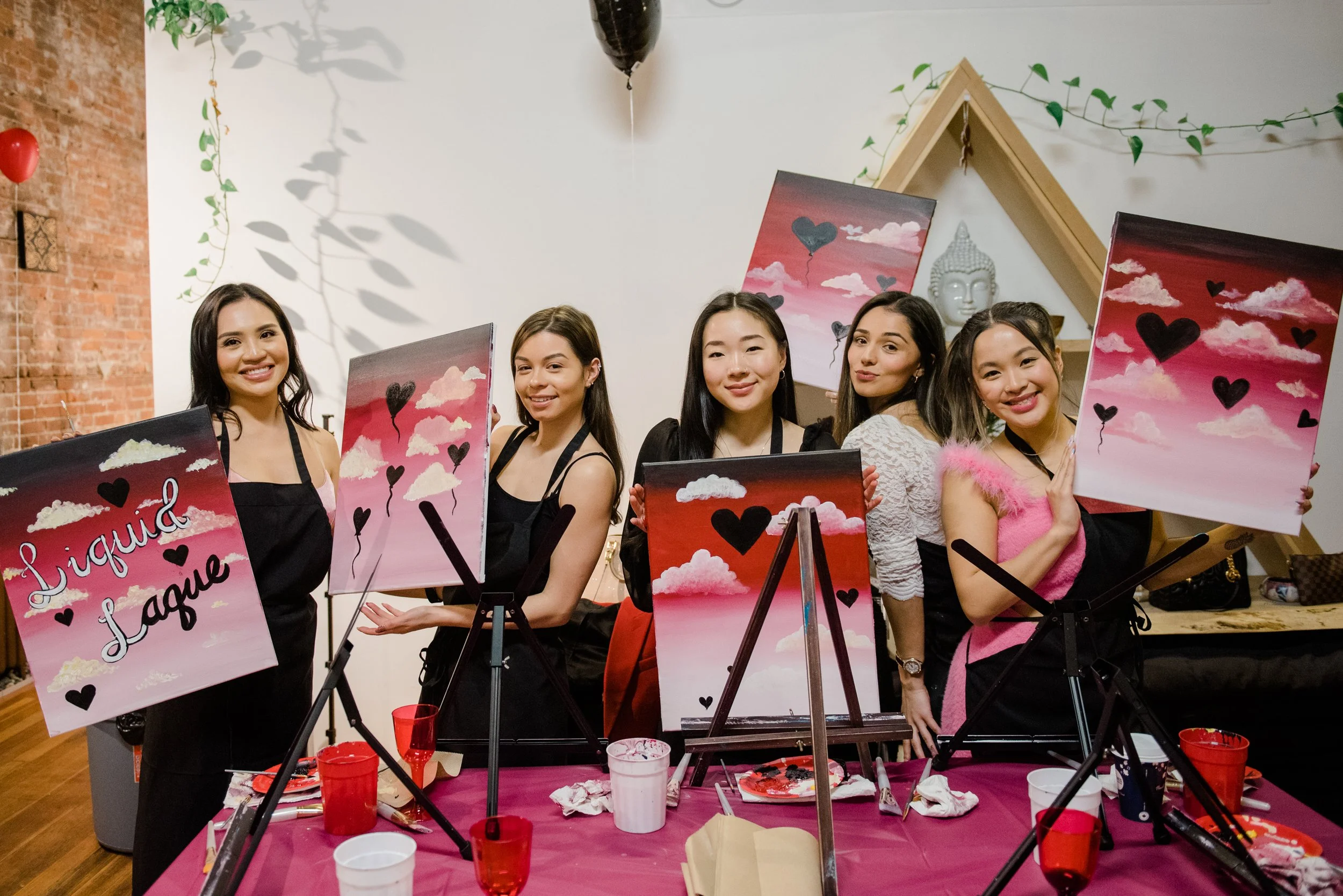 Five women at a painting event, holding paintings with pink sky, clouds, and black heart-shaped balloons, standing behind a table with painting supplies, in a decorated indoor space.