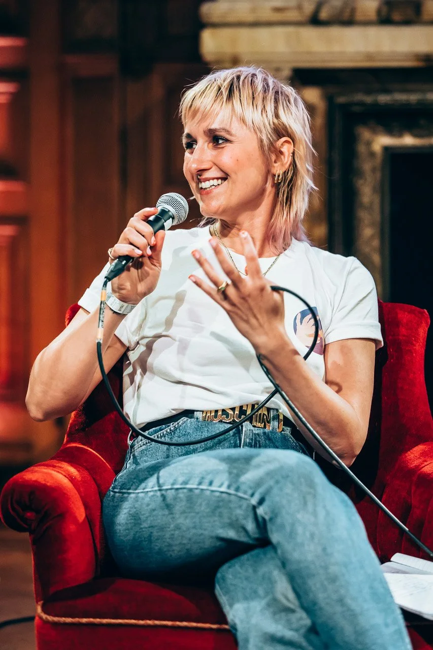 Maxi Meissner at SMUT FEST at De Studio in Antwerp, wearing a white t-shirt and jeans, sitting on a red chair, smiling and holding a microphone.
