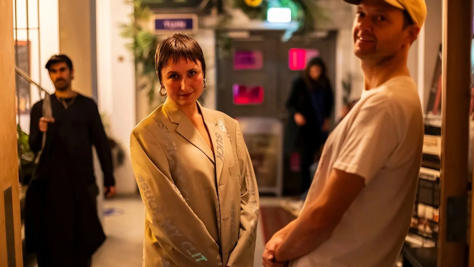Two people standing and facing each other in a dimly lit indoor space at LAB111 in Amsterdam, with a woman in a beige blazer and a man in a beige T-shirt, and two other individuals in the background, one holding a large knife.