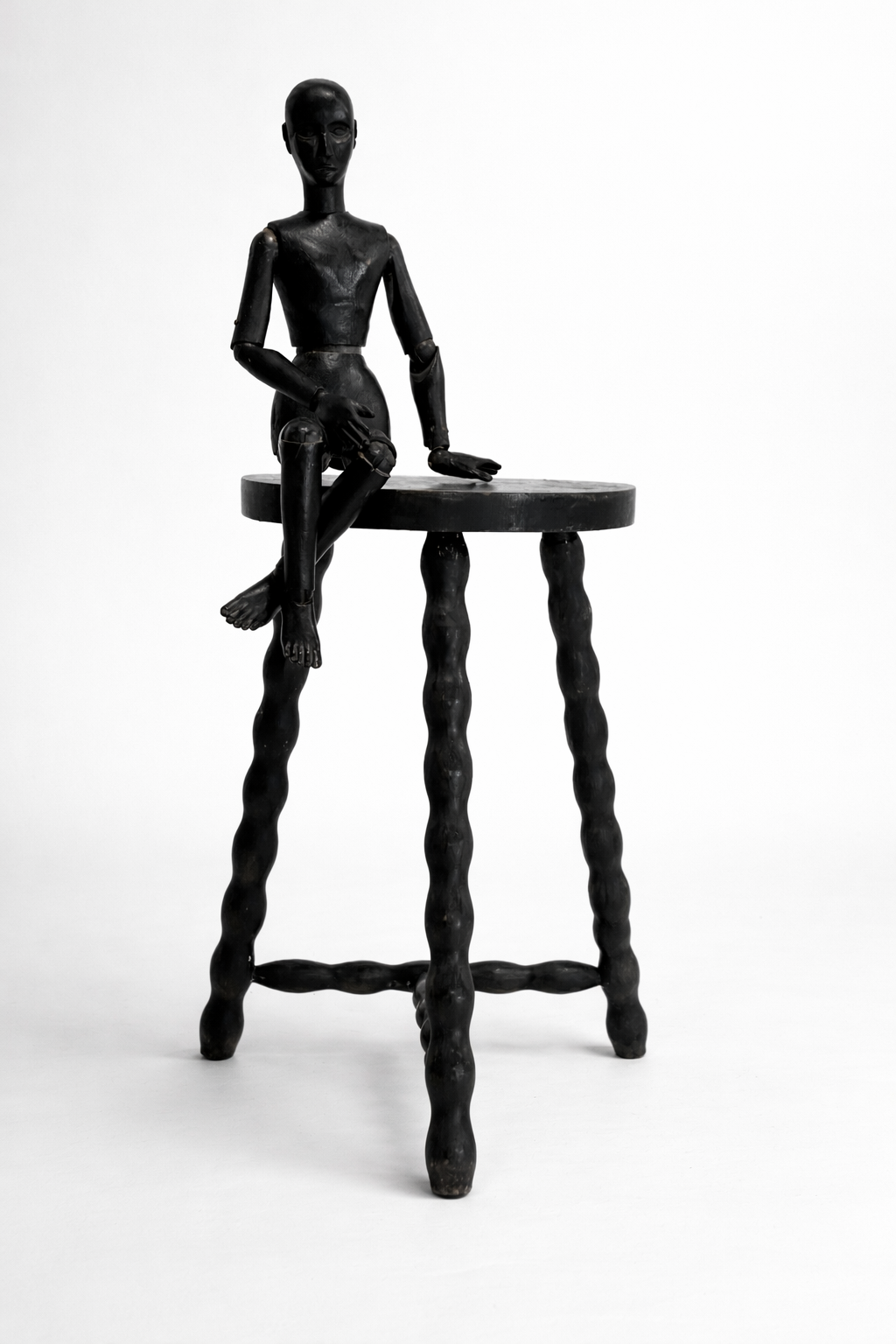 A black wooden mannequin sitting on the edge of a round table with twisted legs, against a plain white background.