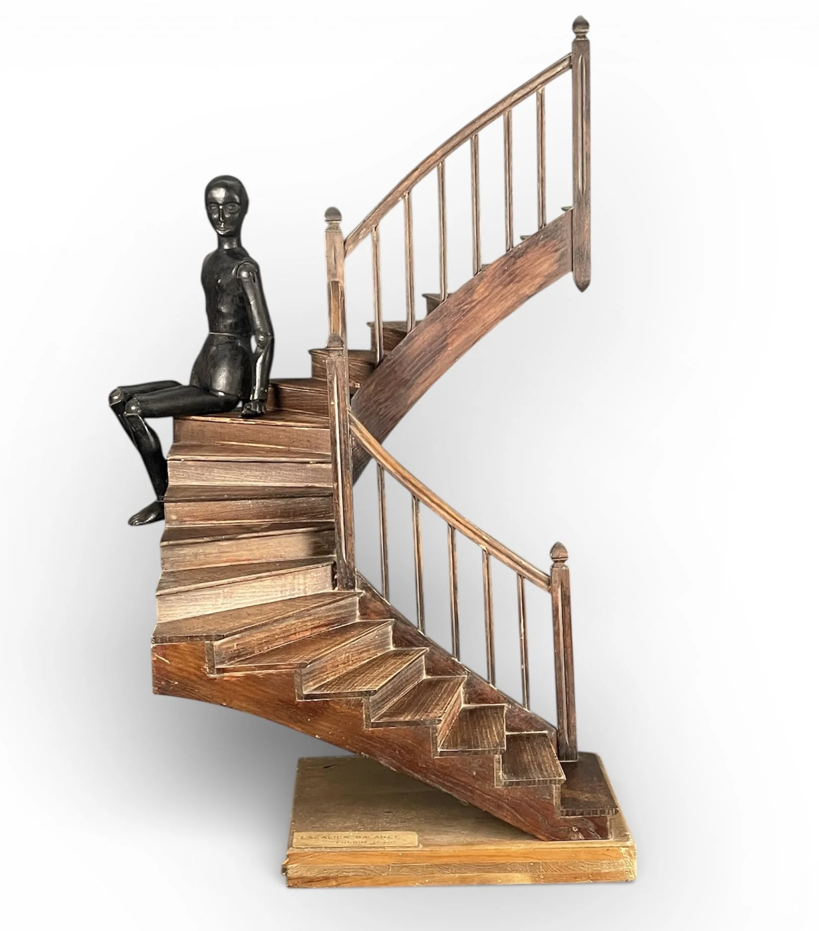 A sculpture of a seated female figure with metallic features, sitting on a wooden staircase with a curved banister, against a plain white background.