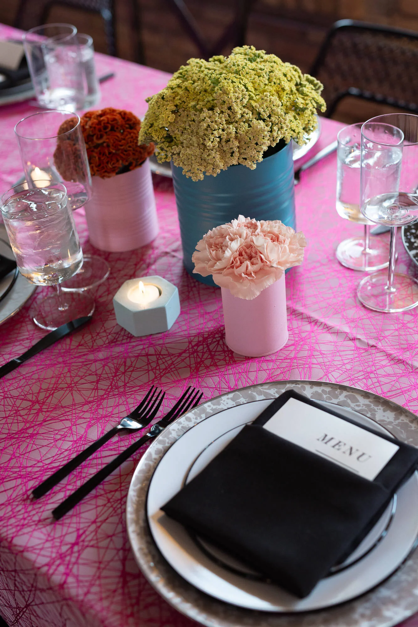  tablescape with pink tablecloth, painted tin can vases with bright floral displays, black silverware and black linens on plates 