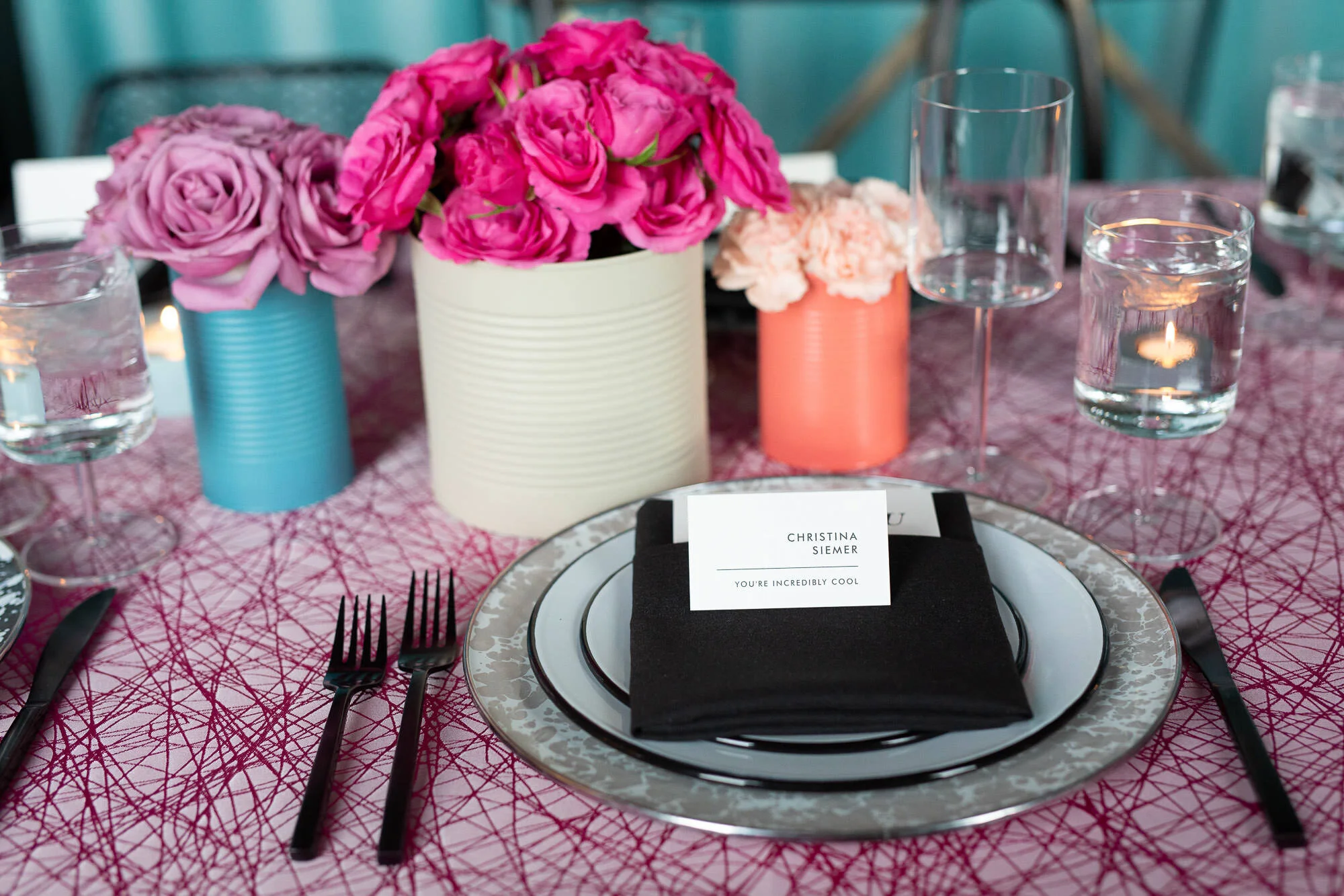  tablescape with pink tablecloth, painted tin can vases with bright floral displays, black silverware and black linens on plates 