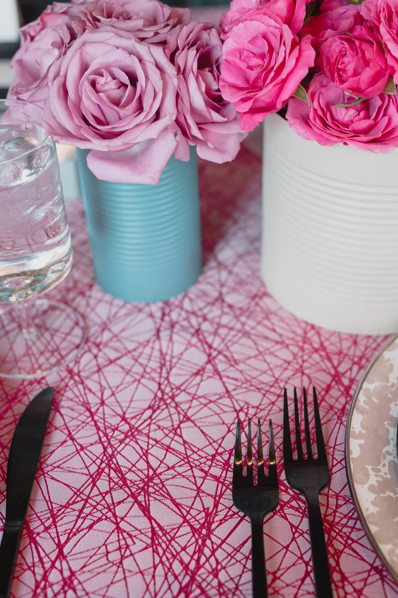  tablescape with pink tablecloth, painted tin can vases with bright floral displays, black silverware and black linens on plates 
