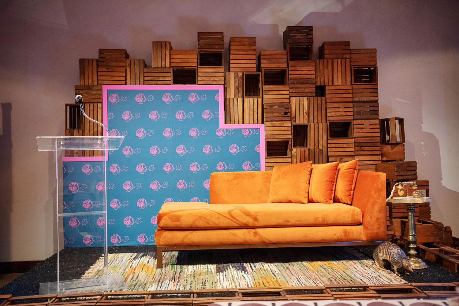  stage display with orange velvet settee, lucite podium, pink and blue floral backdrop, and wooden crates 