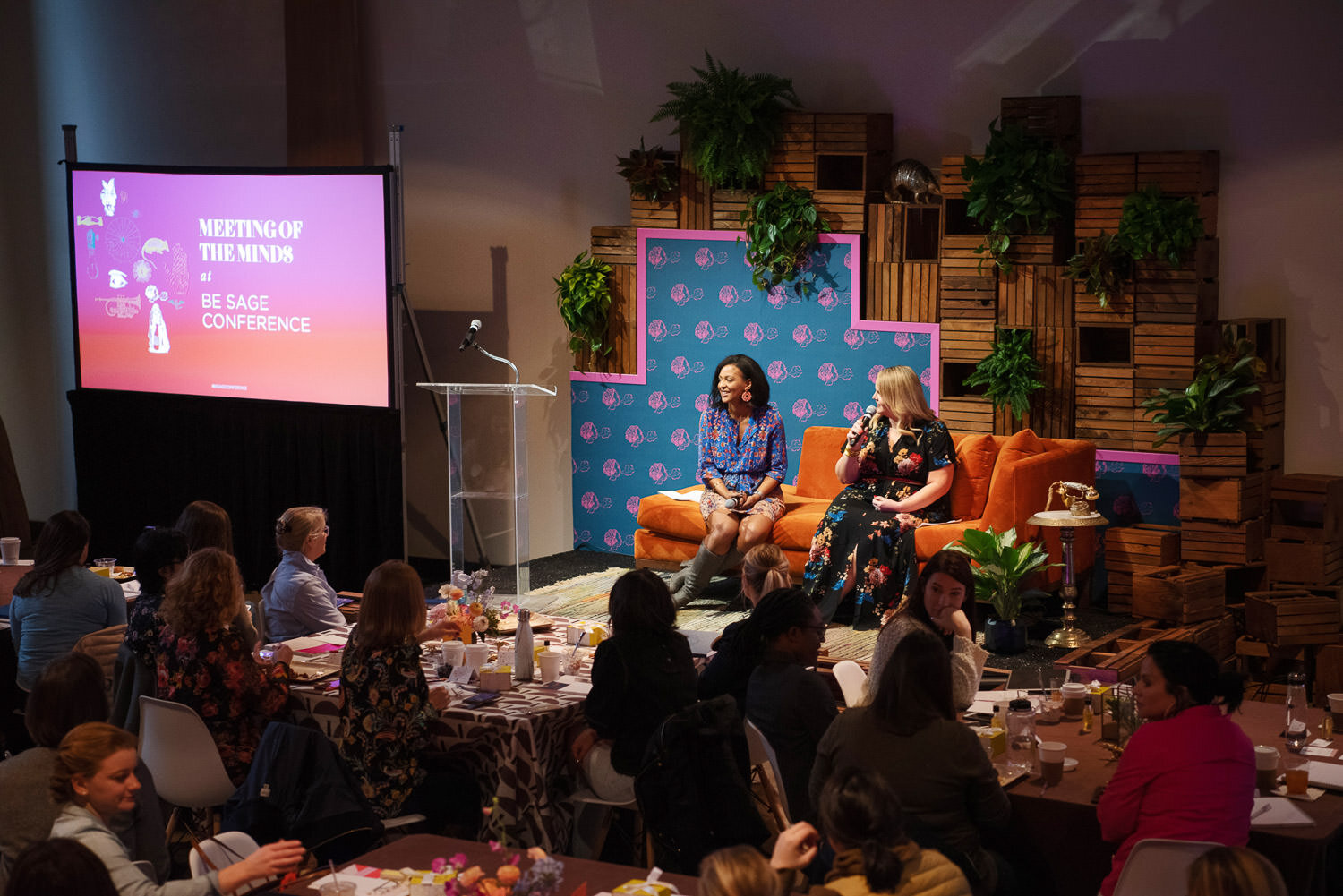  stage display with orange velvet settee, lucite podium, pink and blue floral backdrop, and wooden crates 