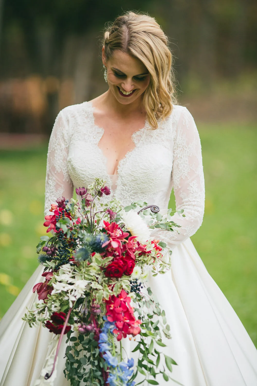  bride in long-sleeved lace dress and large rustic red white and blue bouquet  looking down and smiling 