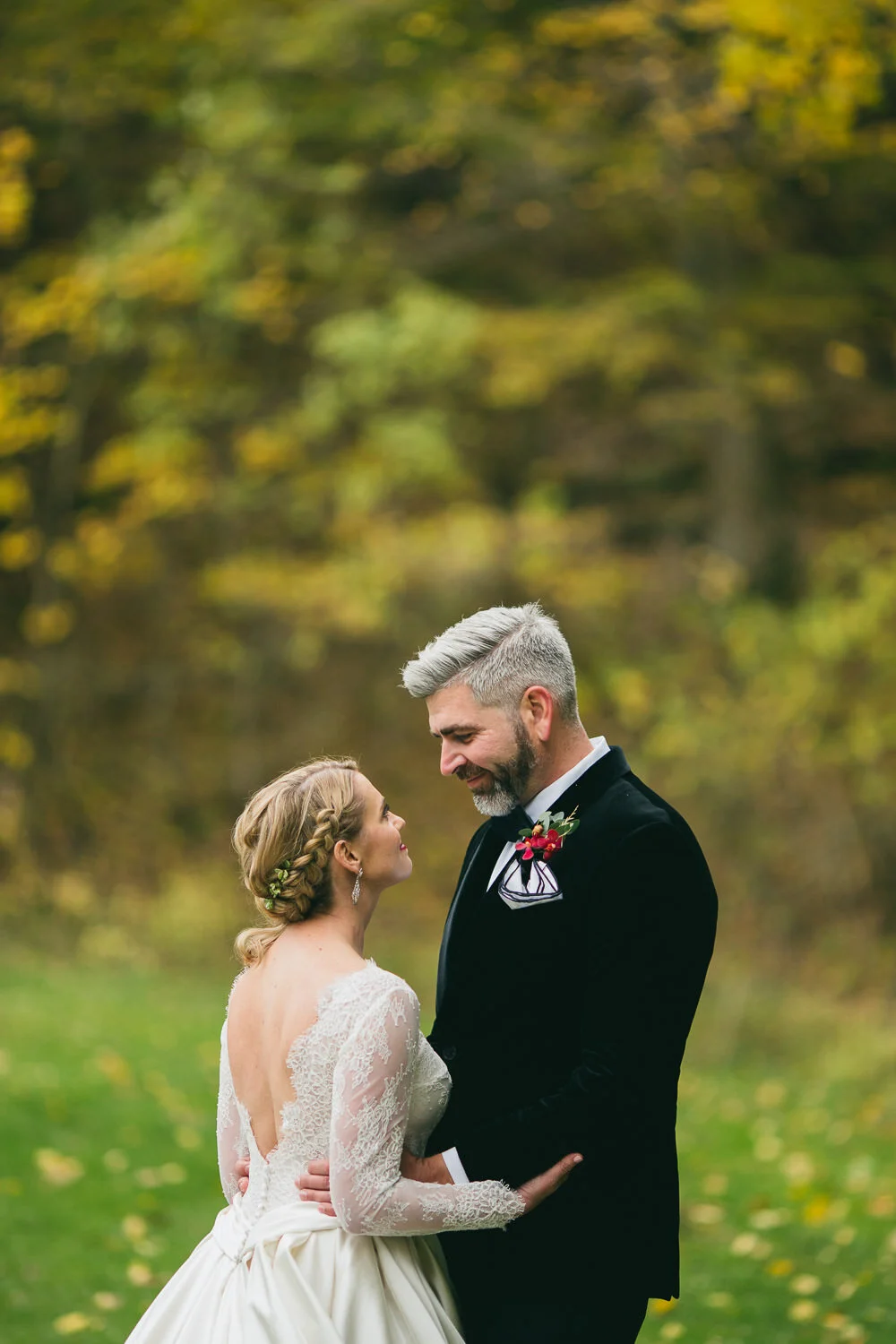  bride in long-sleeved lace dress with plunging low back and braided blonde hair smiles up at salt and pepper haired groom in black tuxedo 