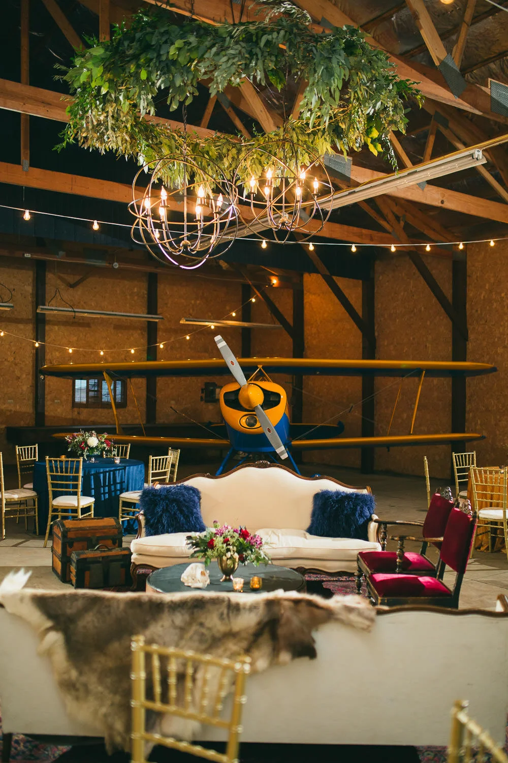  Airplane hangar reception with a yellow and blue vintage airplane, lounge furniture and green floral ceiling installation 