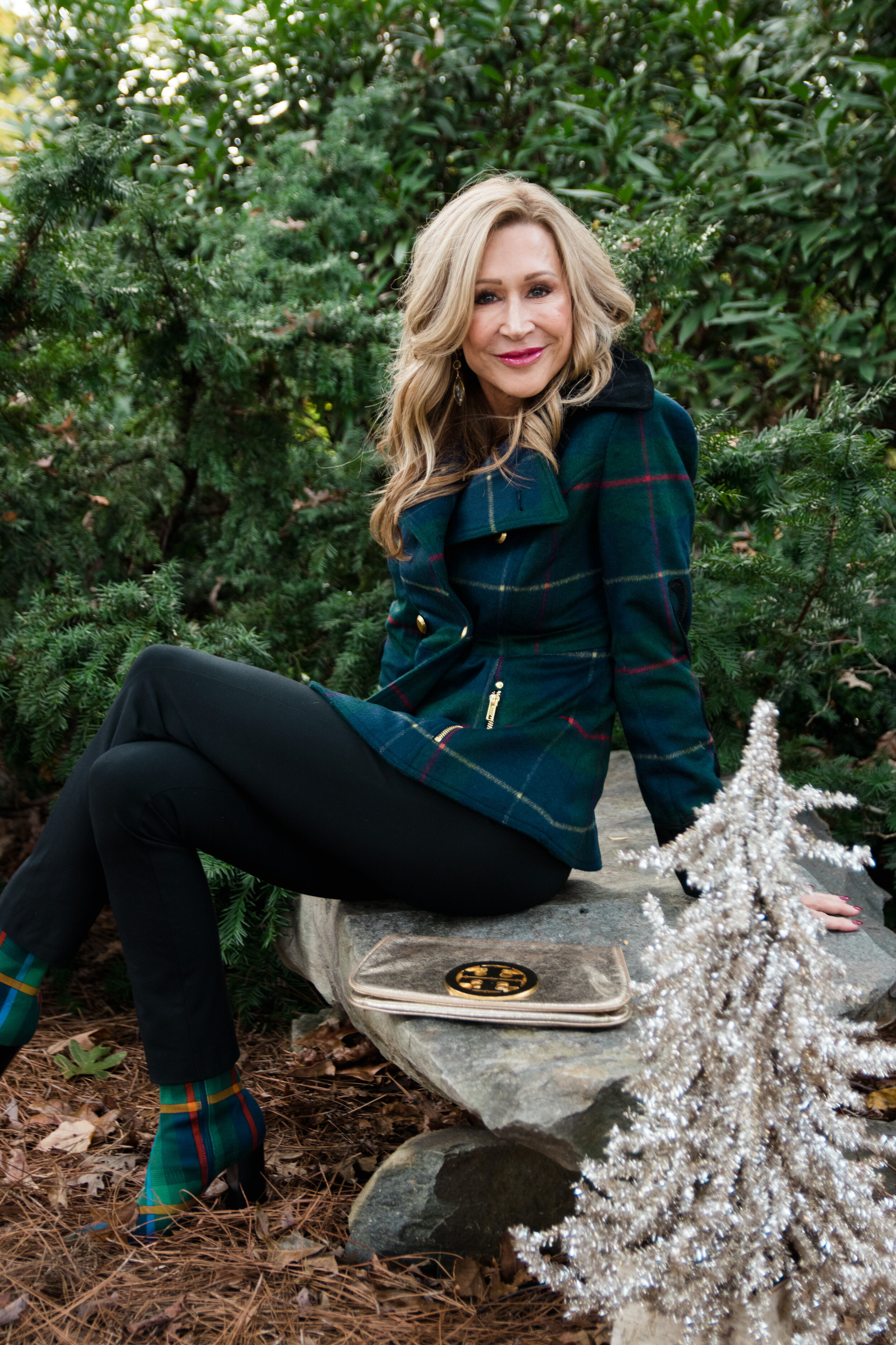 A woman with blonde hair sitting outdoors on a stone bench surrounded by green bushes and trees, wearing a plaid jacket and matching plaid boots, with a small silver Christmas tree and a beige clutch on the bench.