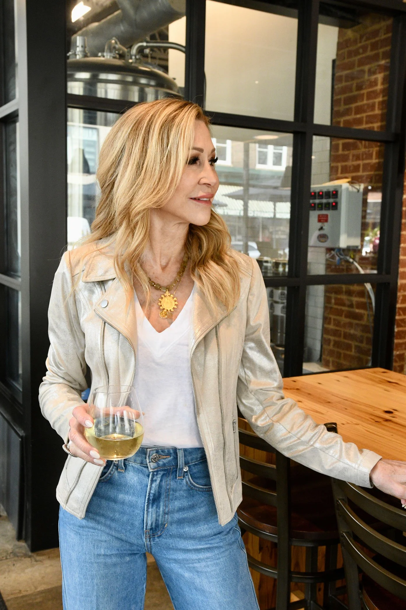 Woman with wavy blonde hair in a beige jacket, white shirt, and blue jeans holding a glass of white wine inside a modern restaurant or cafe.