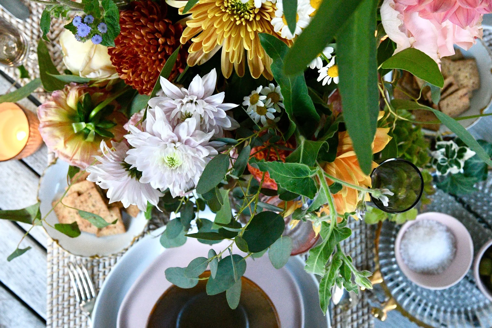 A top-down view of a floral centerpiece with various flowers and greenery on a table set with cups, plates, a candle, and snacks.