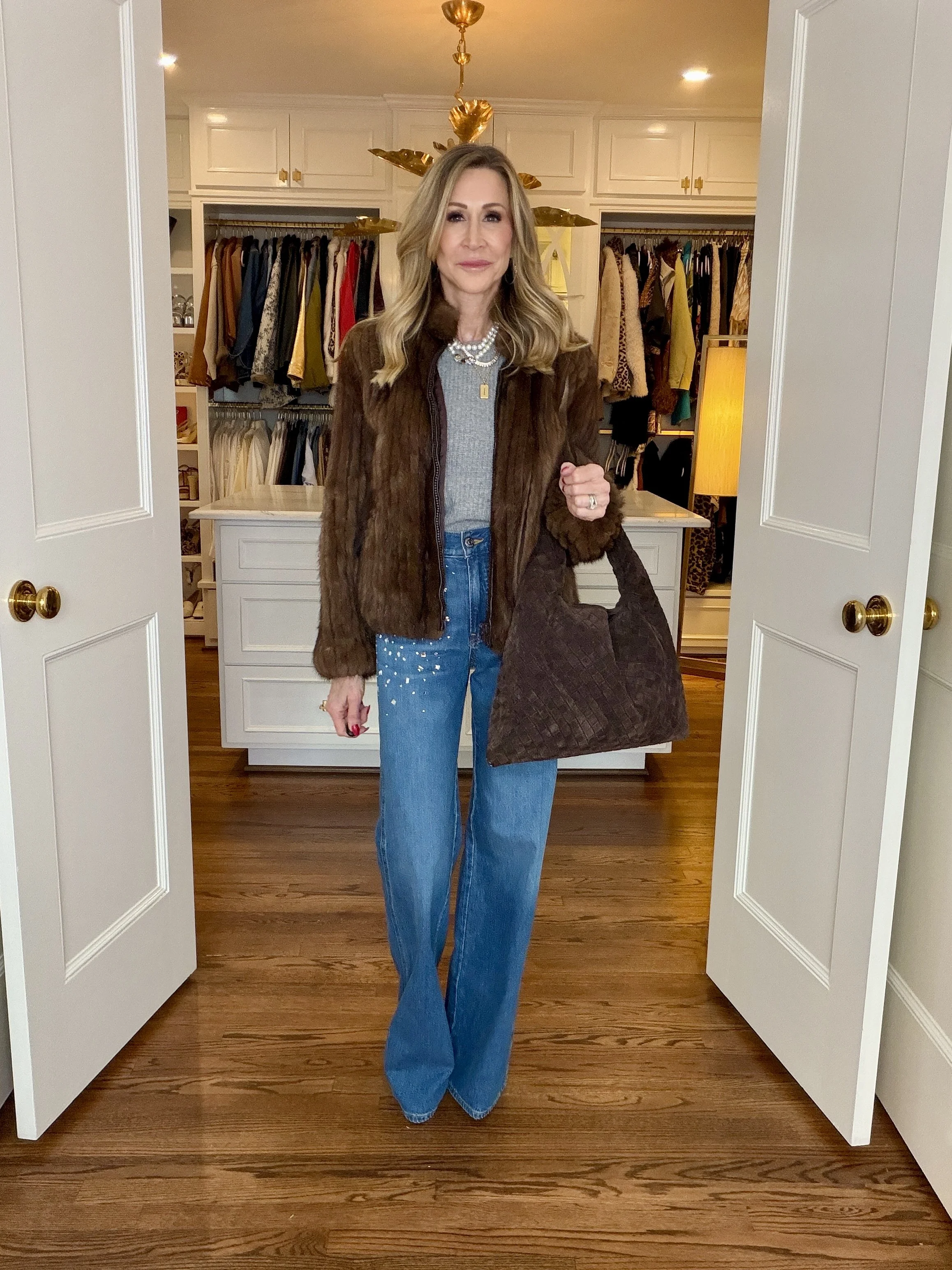 Woman wearing a brown fur coat, layered necklaces, grey sweater, jeans with rhinestones, a brown bag and brown suede boots