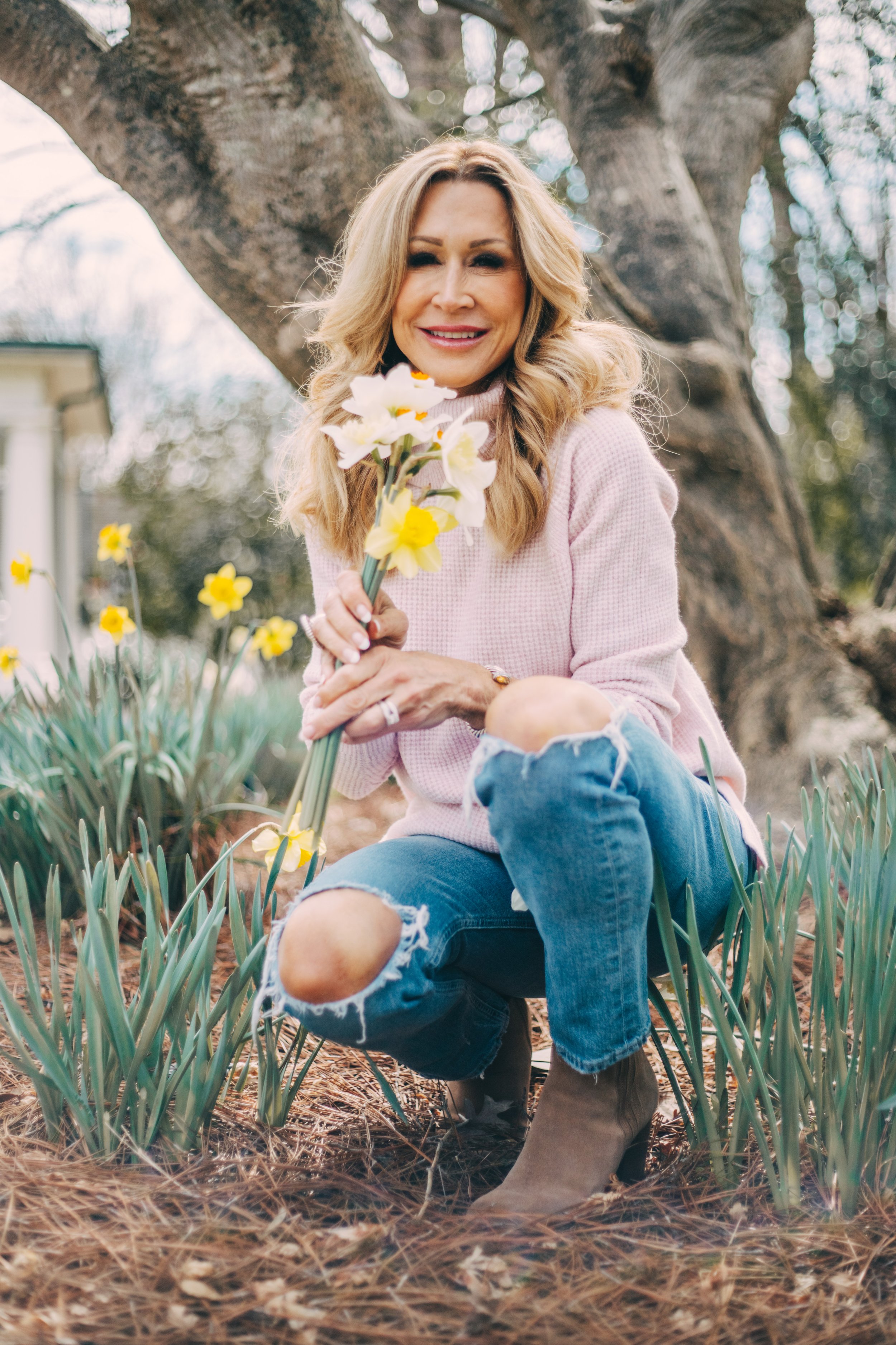 A smiling woman with wavy blonde hair, wearing a pink sweater, ripped jeans, and brown boots, kneeling in a garden holding a bouquet of white and yellow flowers.