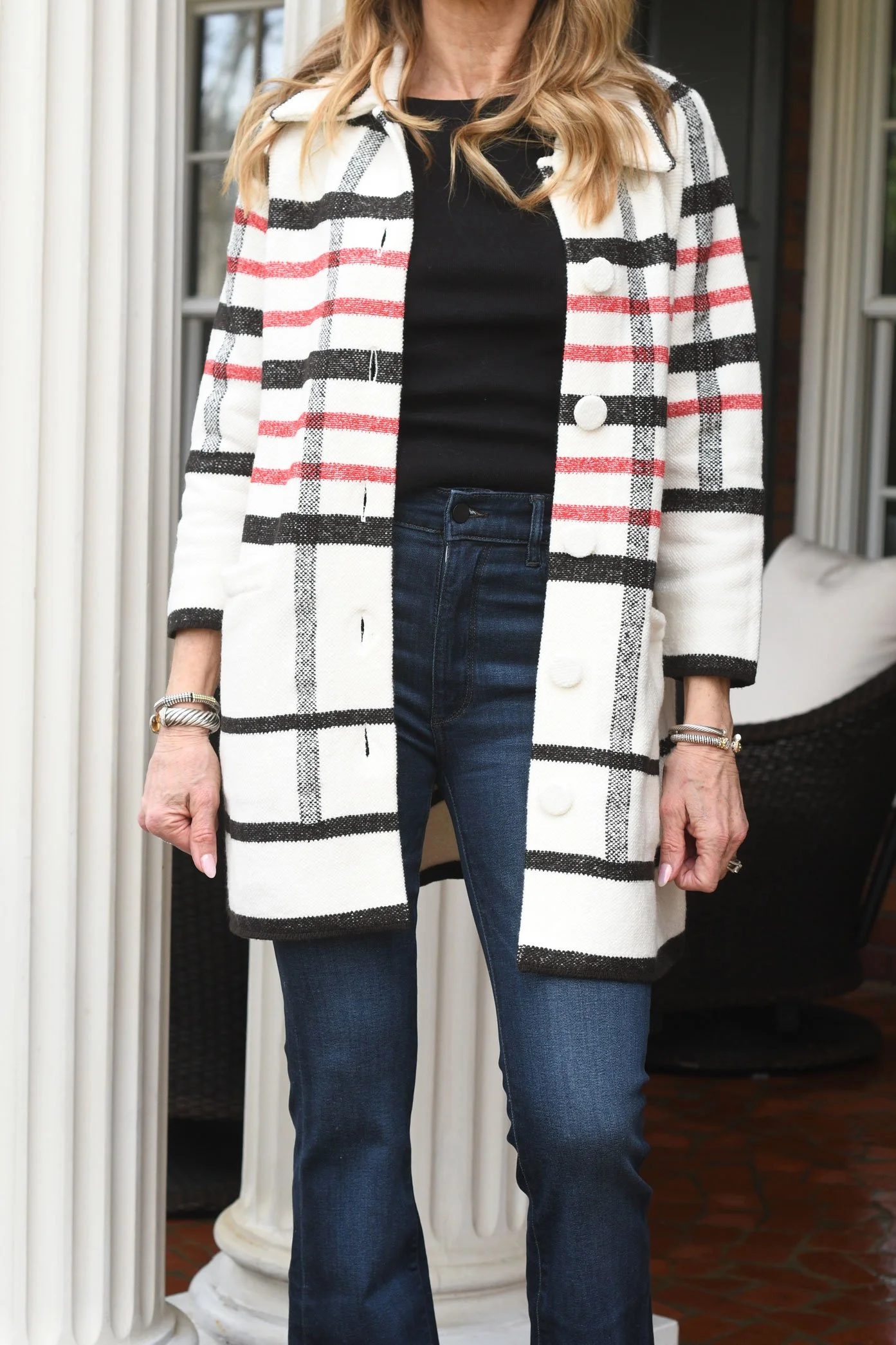 A woman is wearing a black top, dark blue jeans, and an open white coat with black and red plaid pattern, standing outdoors near columns and a wicker chair.