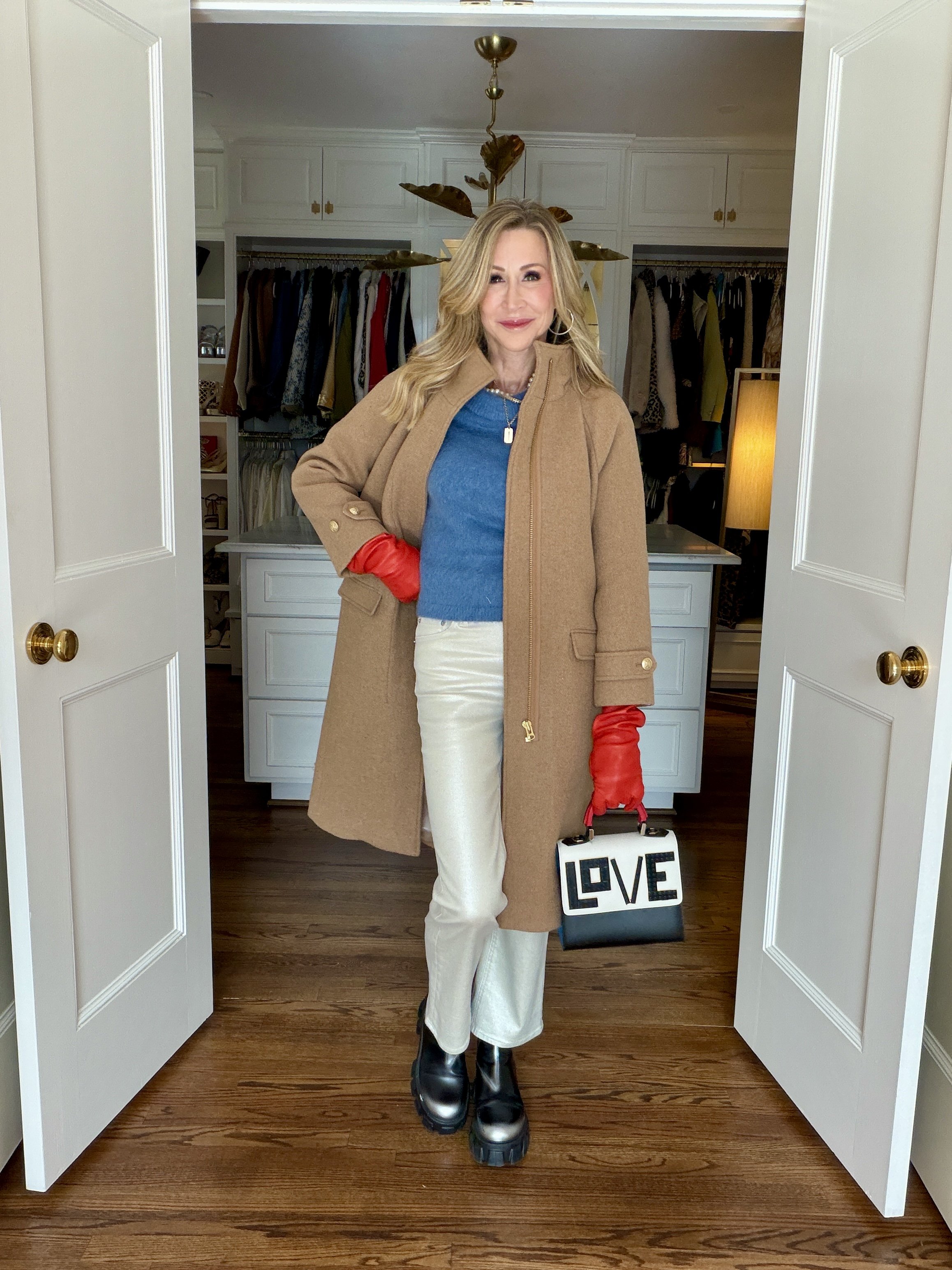 Woman wearing off white jeans, a blue sweater, red leather gloves, lug sole booties, a camel coat and a bag that has the word LOVE written in legos