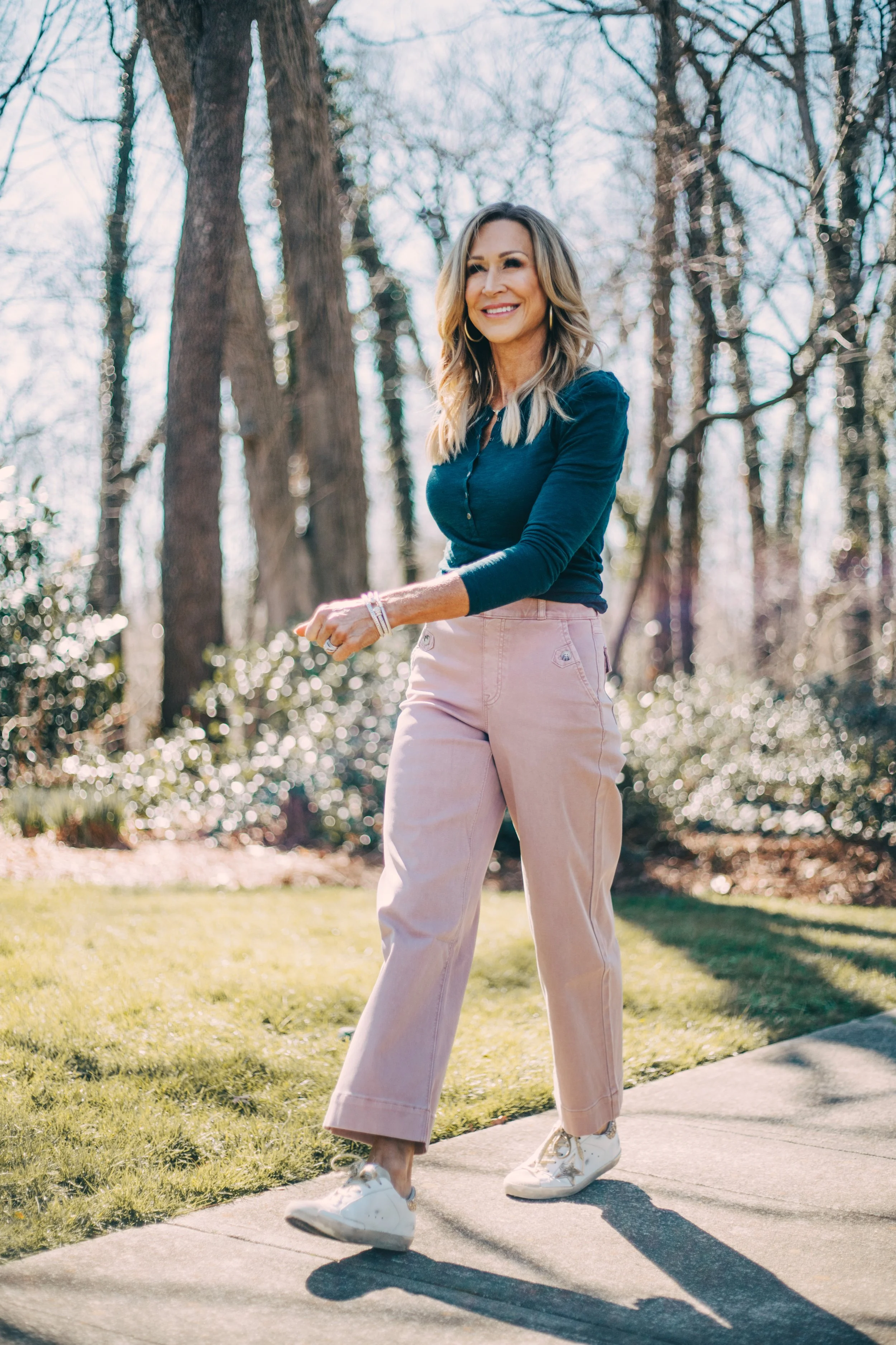 A woman with blonde hair smiling and walking outdoors on a sunny day in a park with trees and bushes in the background, wearing a blue top, light pink pants, and white sneakers.