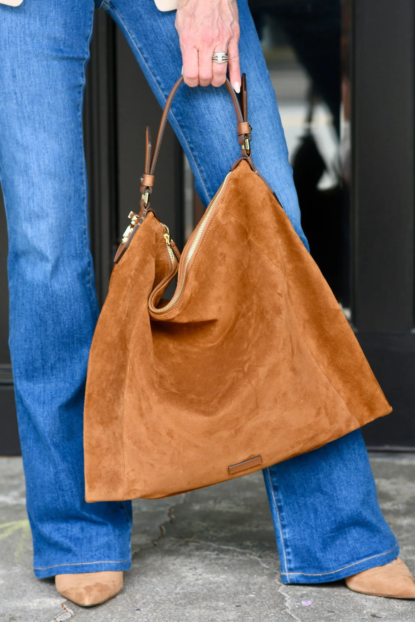 A person holding a large brown suede handbag with a small matching pouch, dressed in blue jeans and tan shoes, standing on a concrete sidewalk.