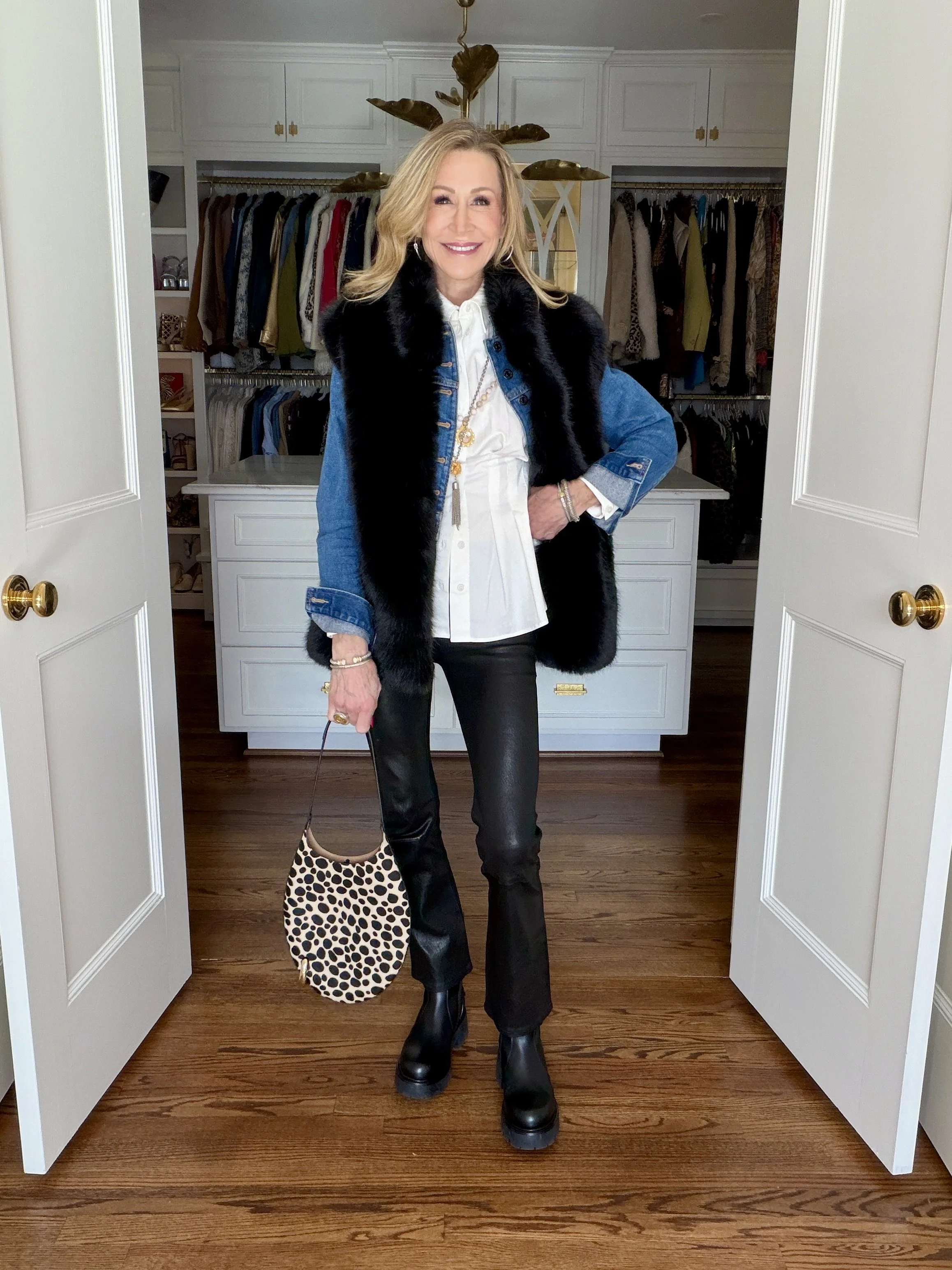 A woman wearing a white blouse, denim jacket, faux fur vest, long necklaces, black leather pants, black boots and a statement bag