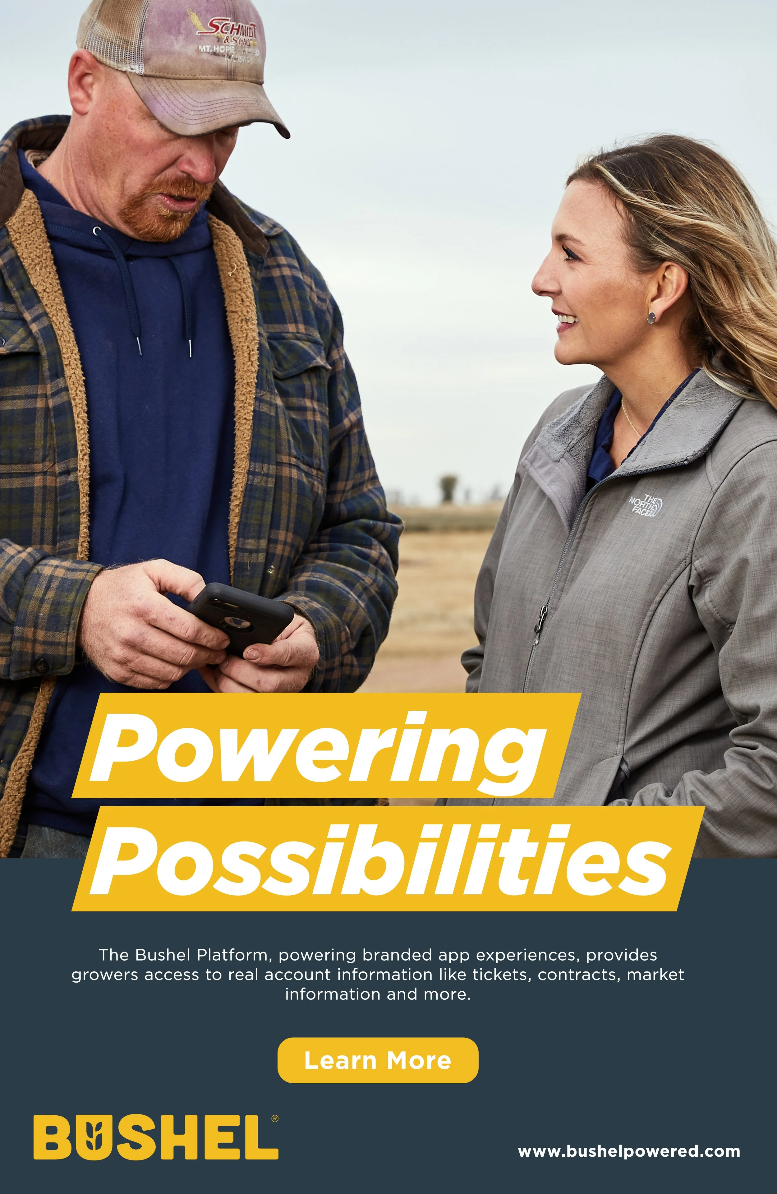 A man and woman outdoors in an open field, both wearing jackets. The man is looking at his phone, and the woman is smiling and looking at him. The text overlay says "Powering Possibilities."